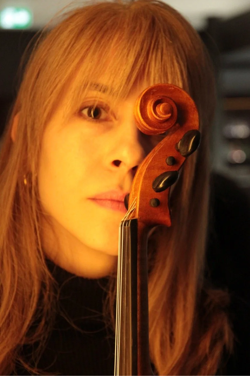 A front profile of a woman, Talia Ramos, holding her violin in an artistic way that shows the scroll hiding her left eye, with a soft warm light.