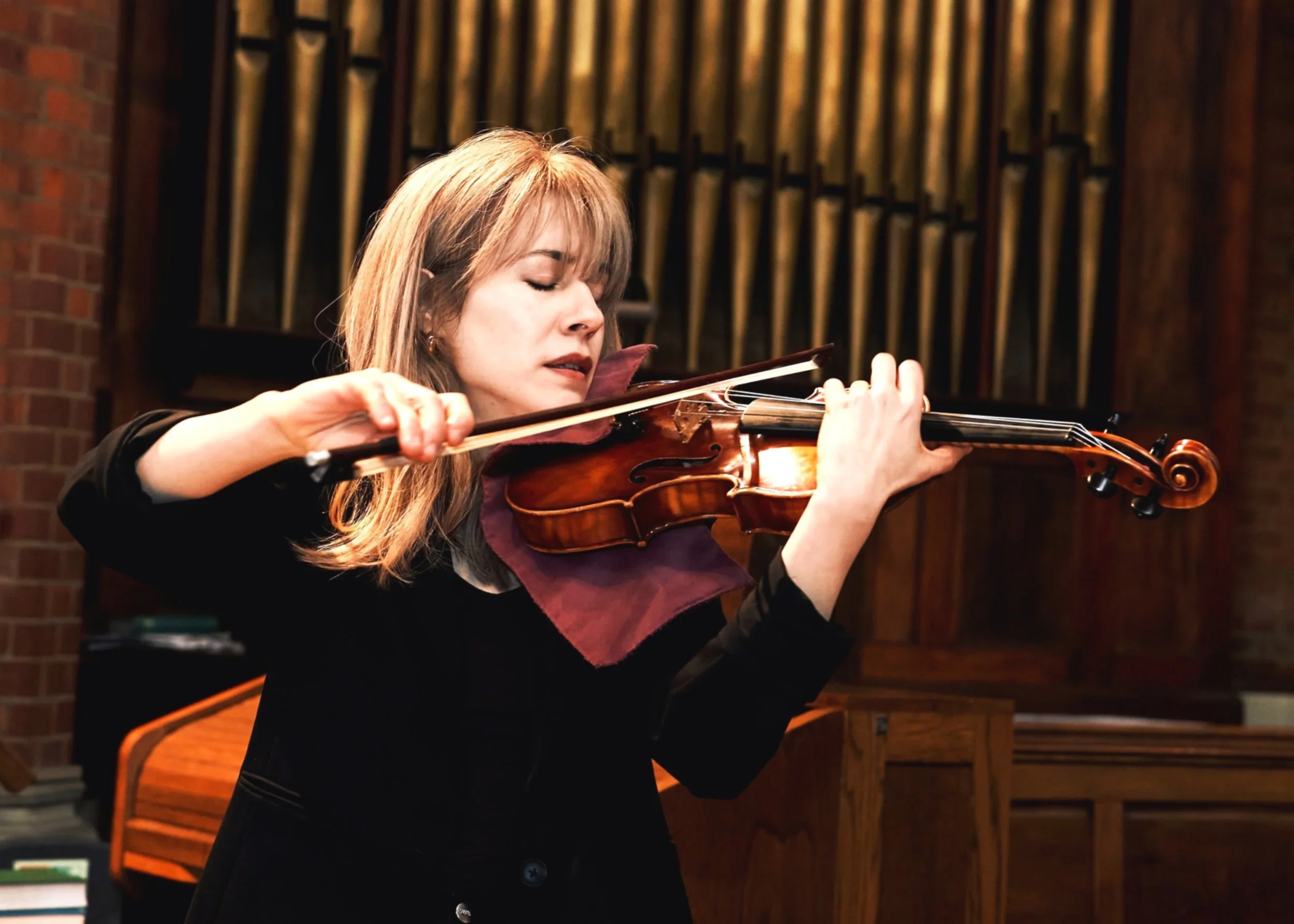 Talia Ramos, with long blonde hair, is playing the violin during a performance in a venue with wooden decor and brick walls.