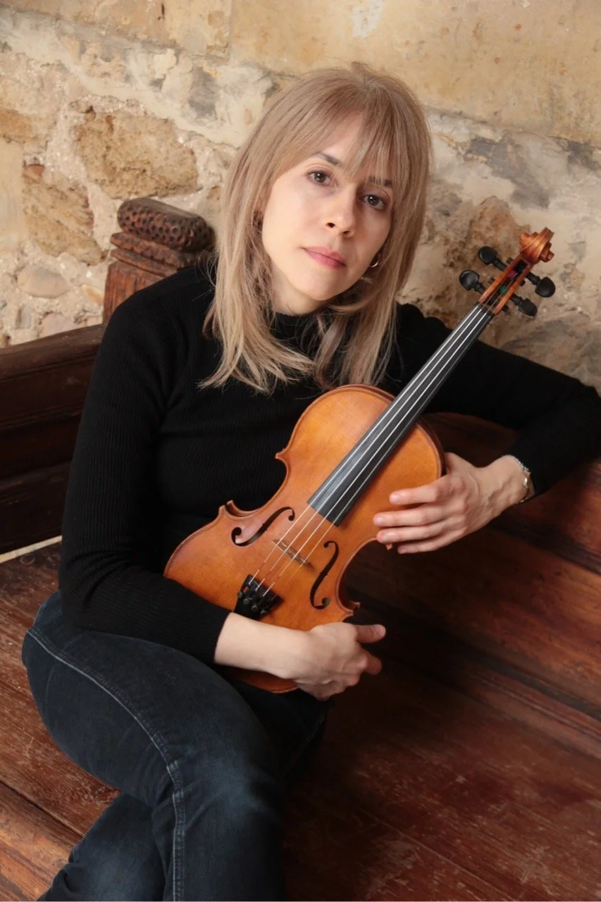 A full body portrait of a woman, Talia Ramos, holding her violin while looking directly at the camera with a soft smile, sitting on a wooden bench against a brick wall.