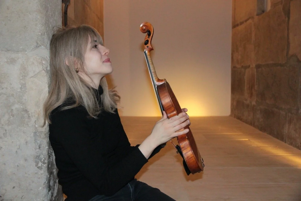 A side portrait of violinist Talia Ramos, holding her violin and smiling against a soft background