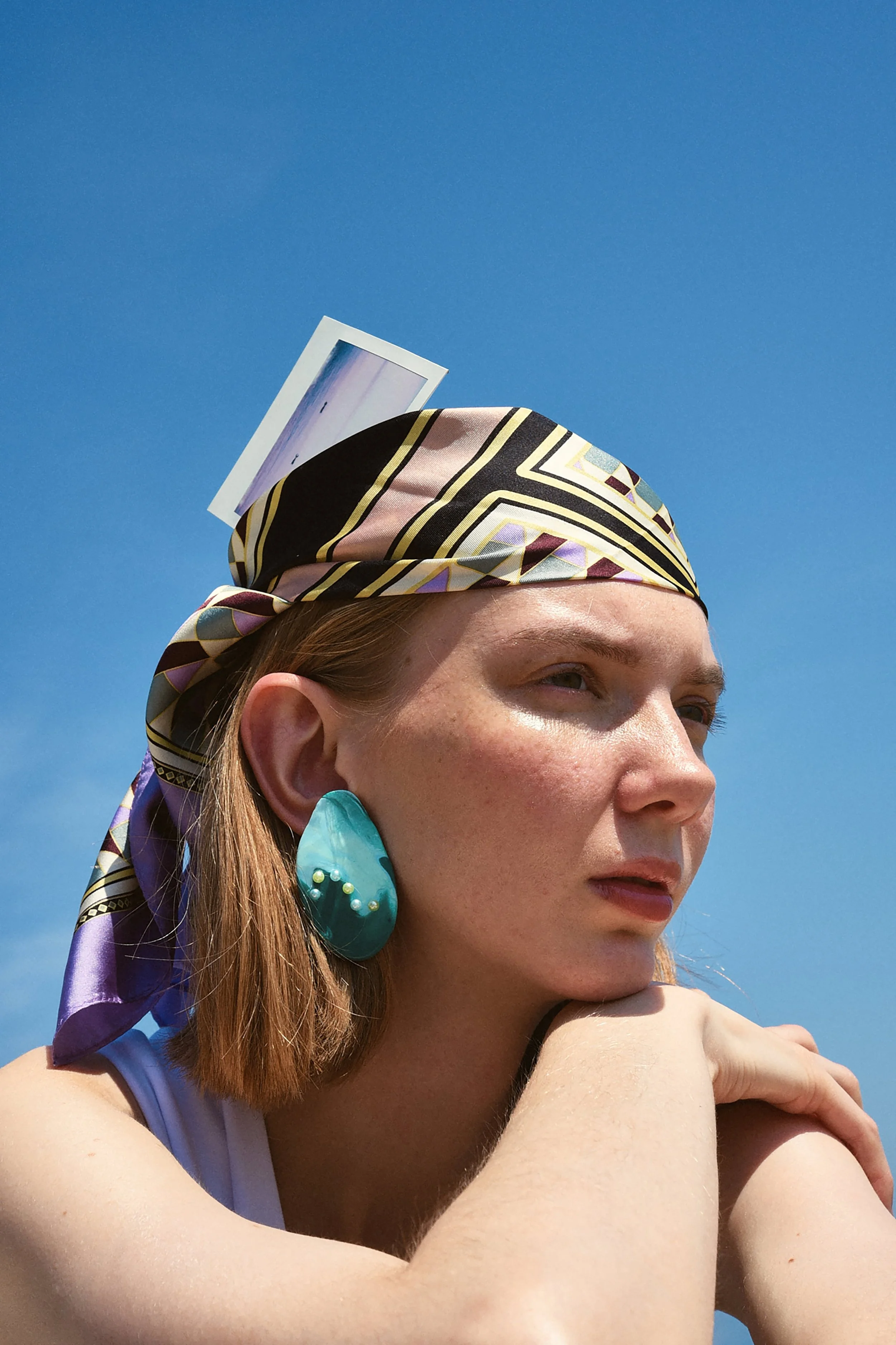 Ragazza con sciarpa colorata e orecchino blu, guarda verso l'orizzonte sotto un cielo azzurro, con una fotografia attaccata alla sciarpa.