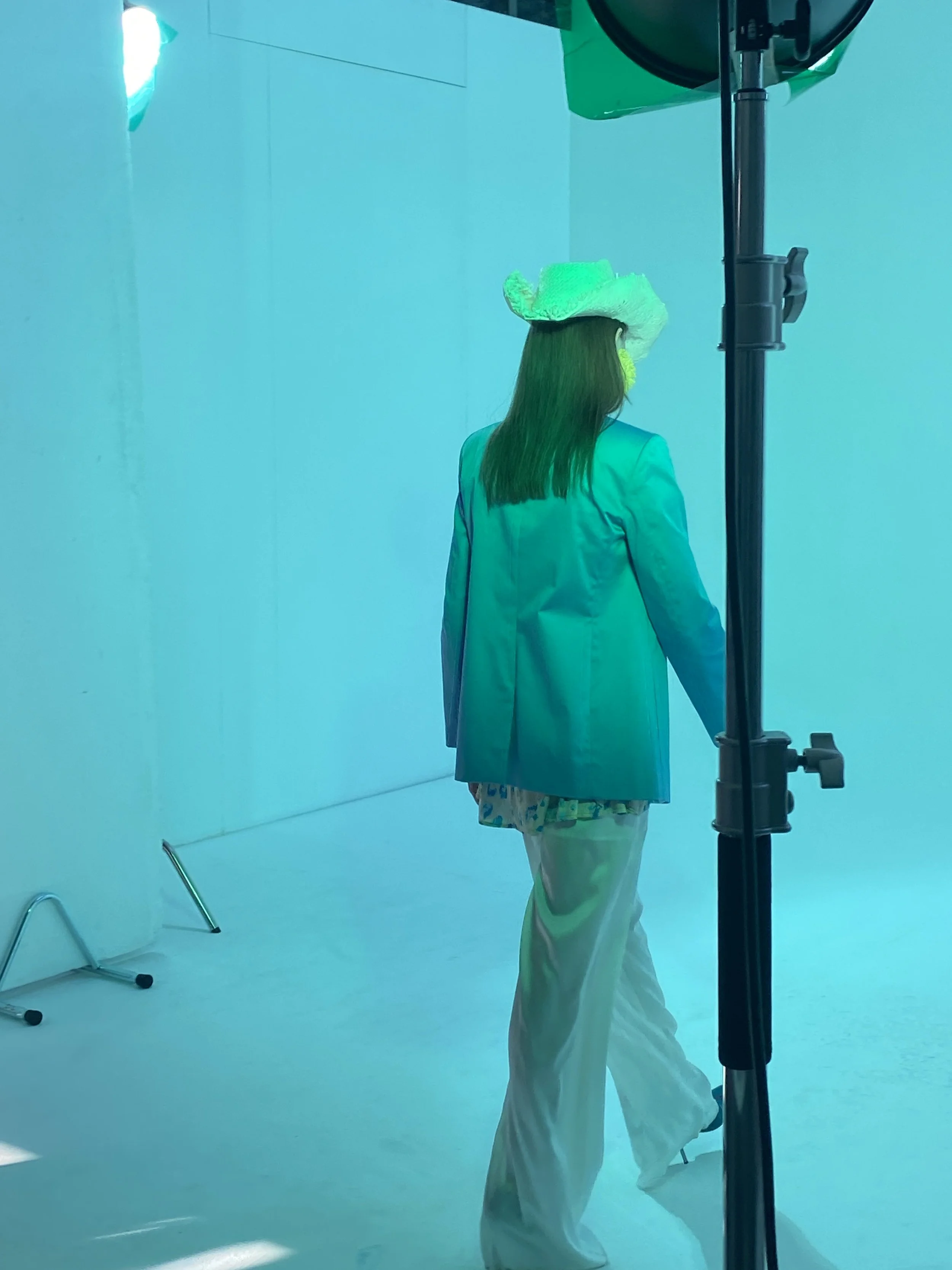 Donna con cappello bianco, abito bianco e pantaloni bianchi, vista di schiena, in uno studio fotografico con illuminazione a colori