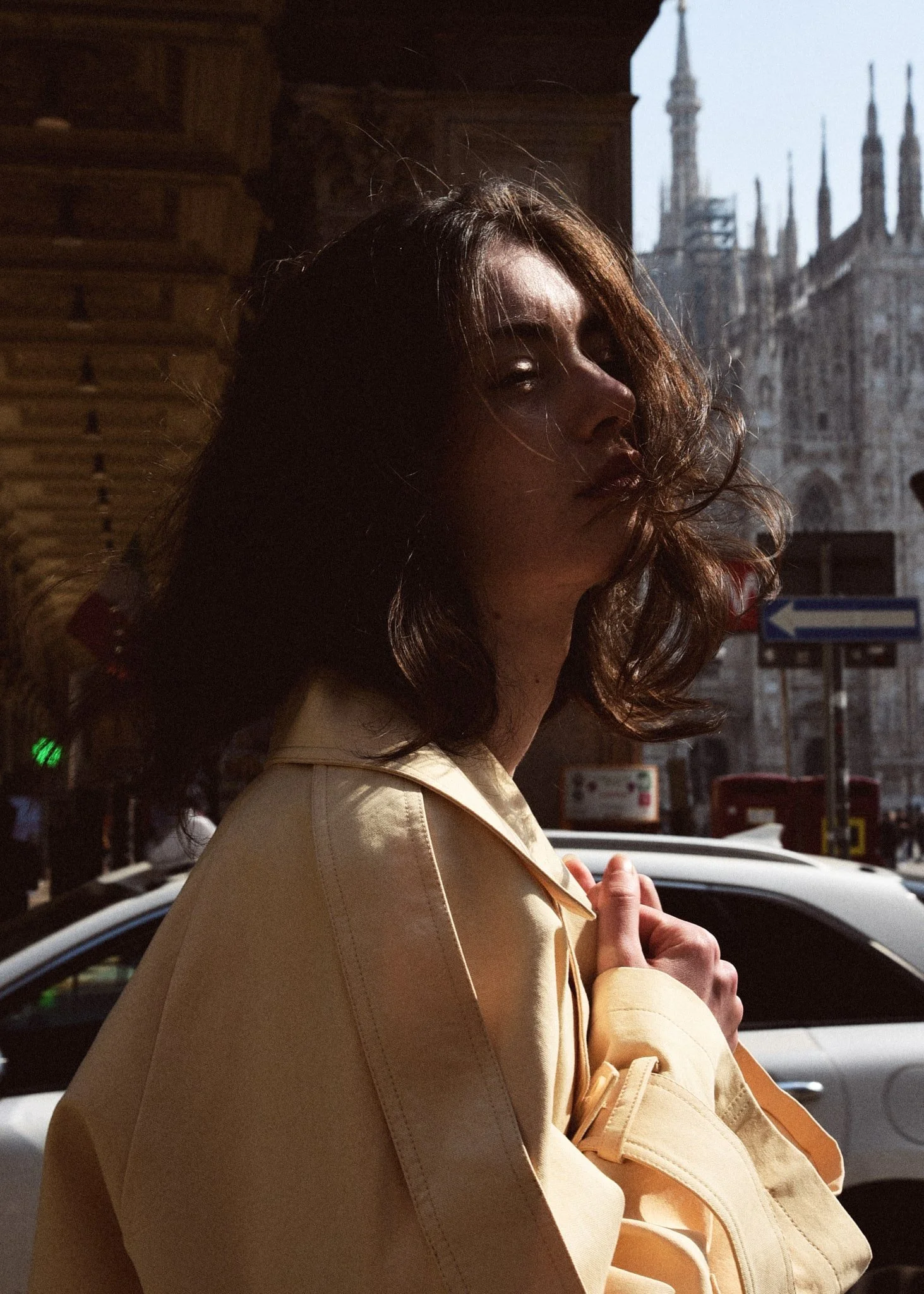 Ragazza con capelli ondulati e cappotto beige, in piedi in strada con edifici storici sullo sfondo.