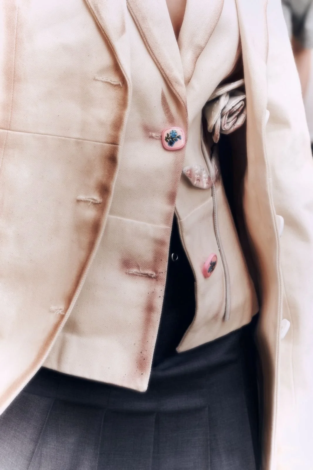 Un close-up di una donna che indossa una giacca beige con bottoni decorativi e una camicia di seta a motivo floreale, con una gonna nera.