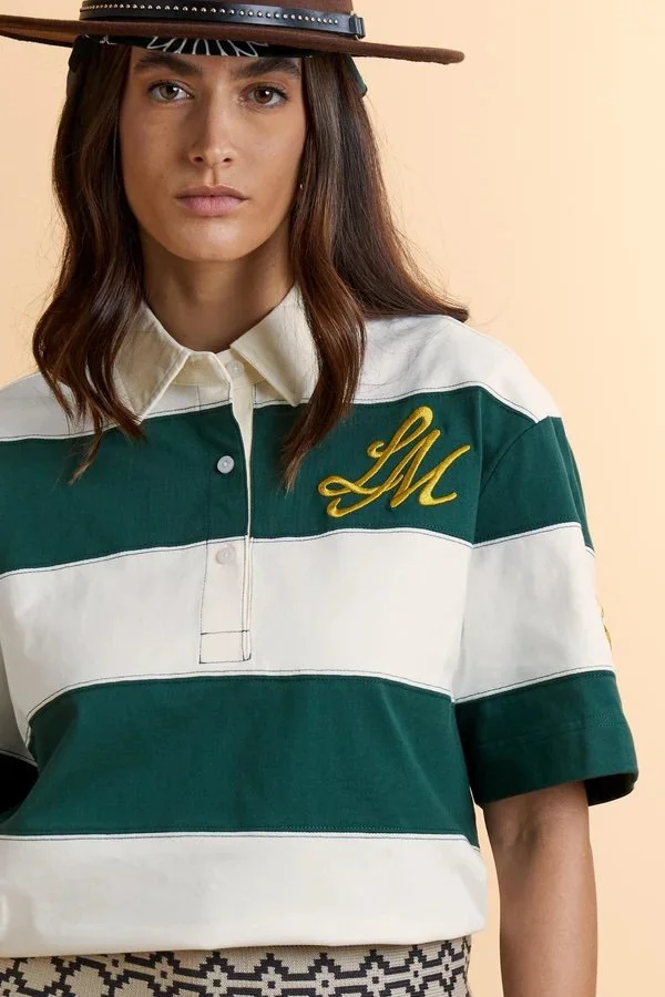 Donna con cappello a secchiello, maglia rugby a righe verde e bianca con ricamo GMS argentato, sfondo beige.
