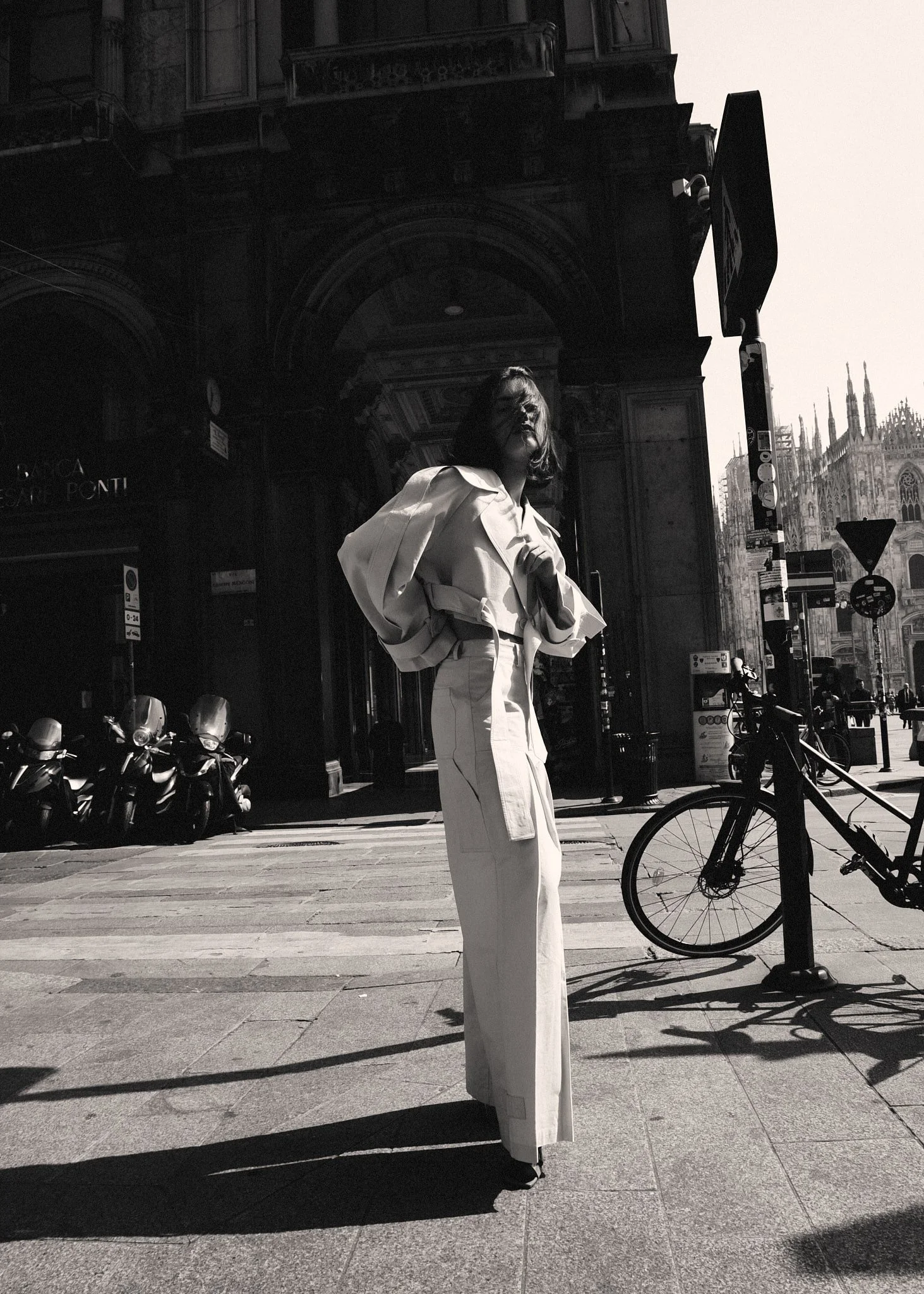 Una donna con abbigliamento chiaro in piedi su un marciapiede nel centro di Milano, con biciclette e motociclette sullo sfondo, e un edificio storico in lontananza, in una foto in bianco e nero.