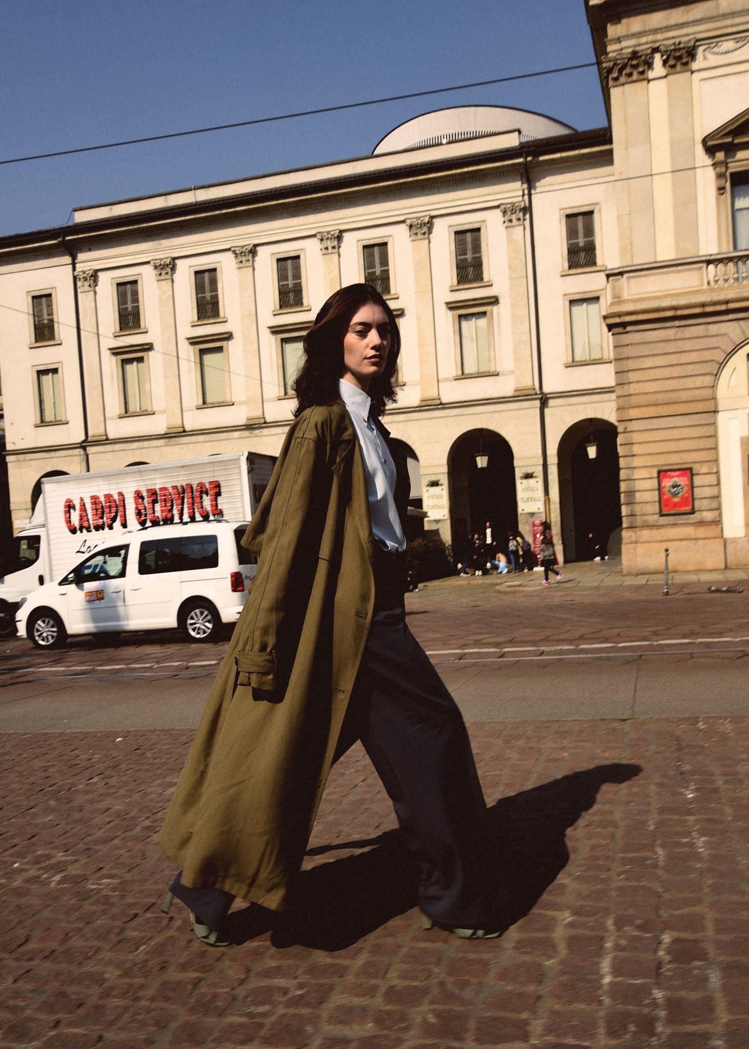 Una donna cammina lungo una strada di pietra con un edificio storico sullo sfondo, un camion con scritto 'Carpi Service' e alcune persone in lontananza.