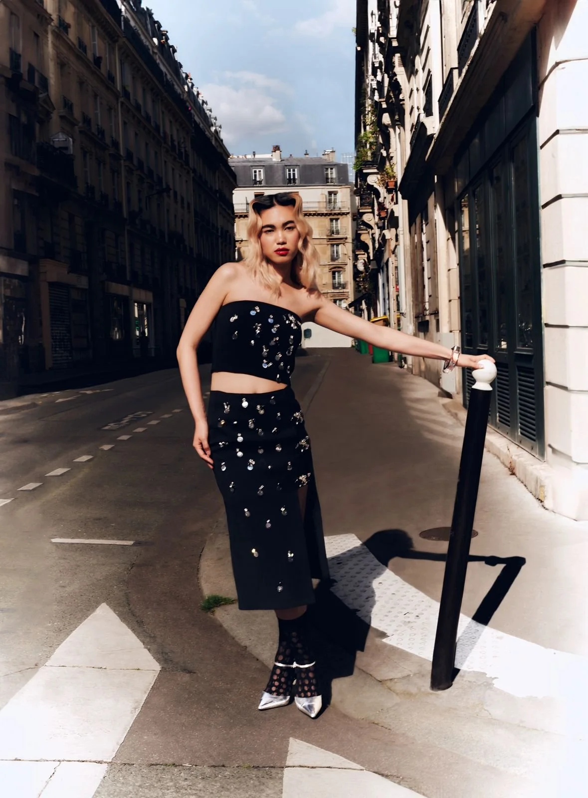 Una donna con capelli biondi, vestita con un abito nero decorato con paillettes, sta in un angolo di strada di Parigi, tenendo una barra di metallo. Sfondo di edifici storici in stile parigino e un cielo nuvoloso.