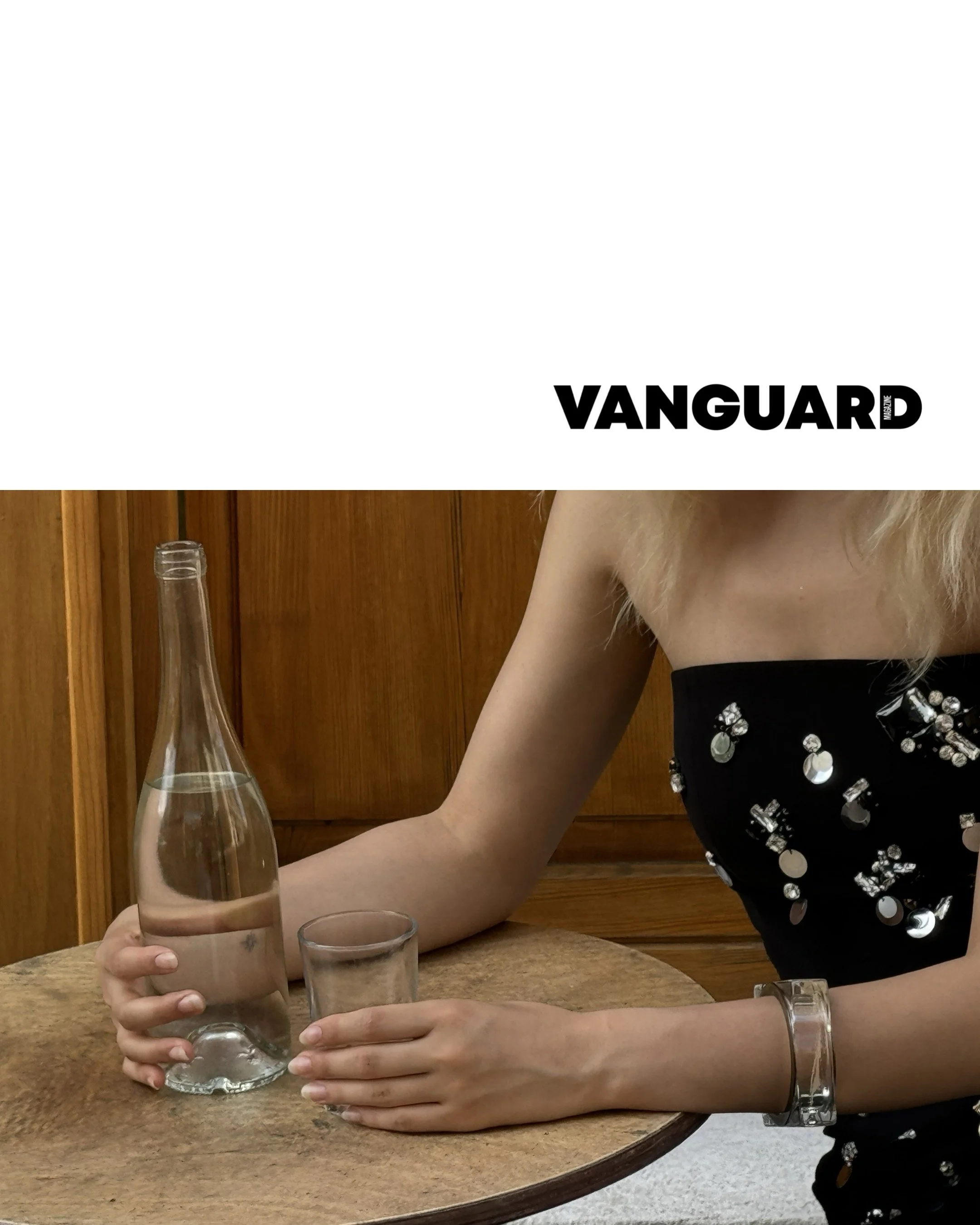 Persona che siede a un tavolo in legno, con una bottiglia di acqua e un bicchiere. Indossa un vestito nero decorato con pietre brillanti, bracciali trasparenti e ha capelli biondi lunghi.