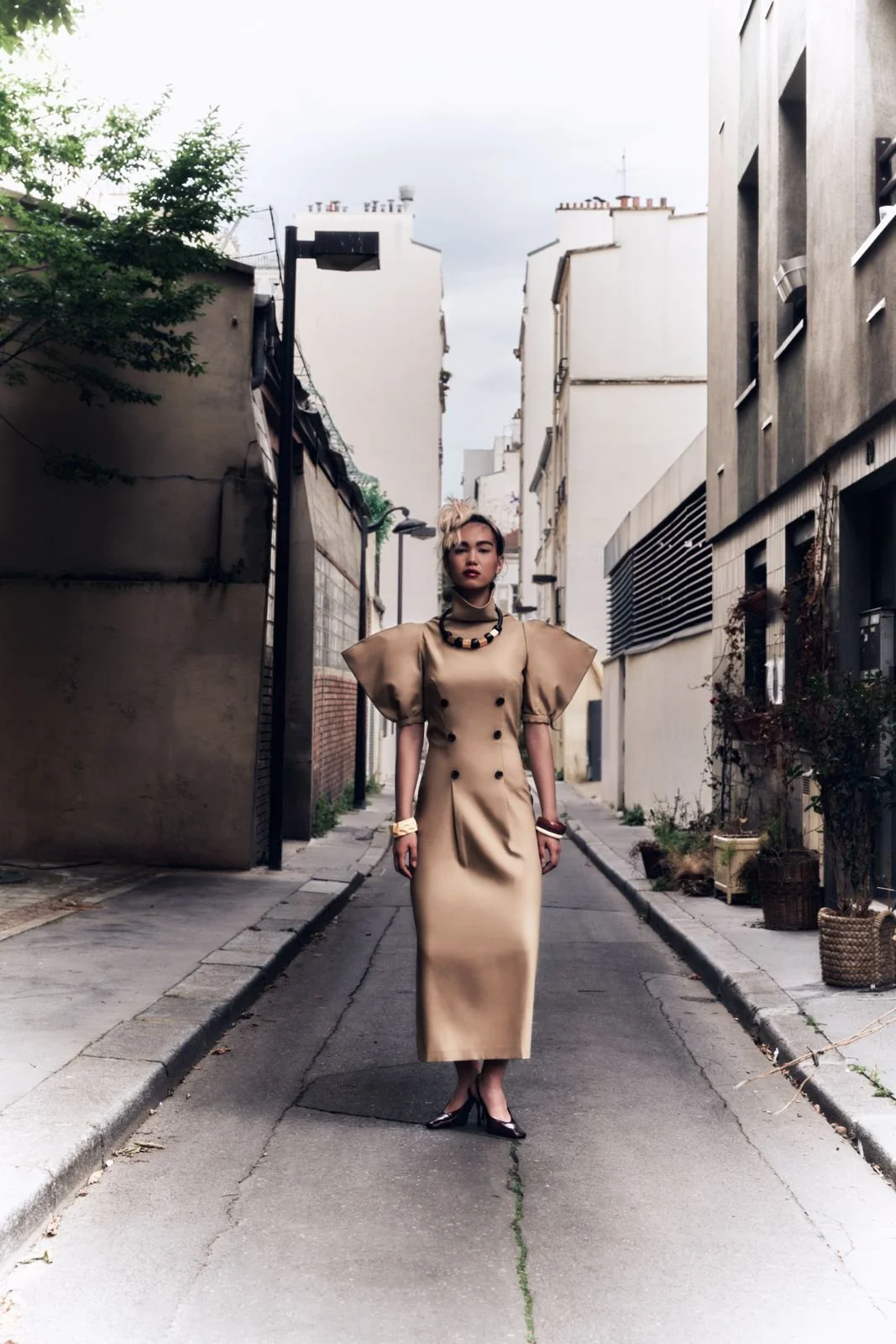 Donna in abito lungo beige con spalle larghe, cintura in vita, sciarpa e accessori neri, cammina in una stradina urbana, con edifici moderni e piante.