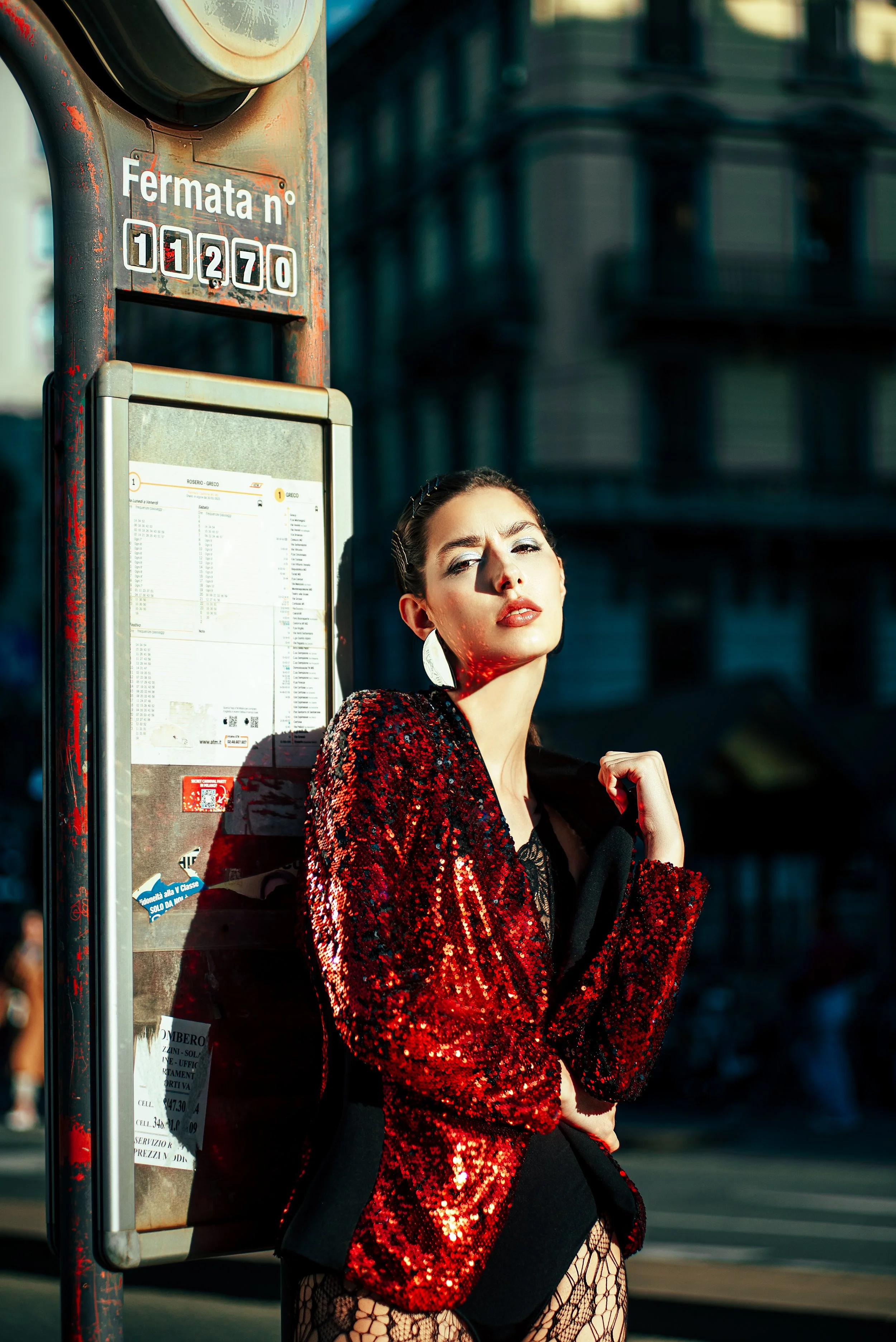 Fermata dell'autobus con una donna vestita elegantemente in posa davanti all'edicola, illuminata dalla luce del tramonto a Milano.