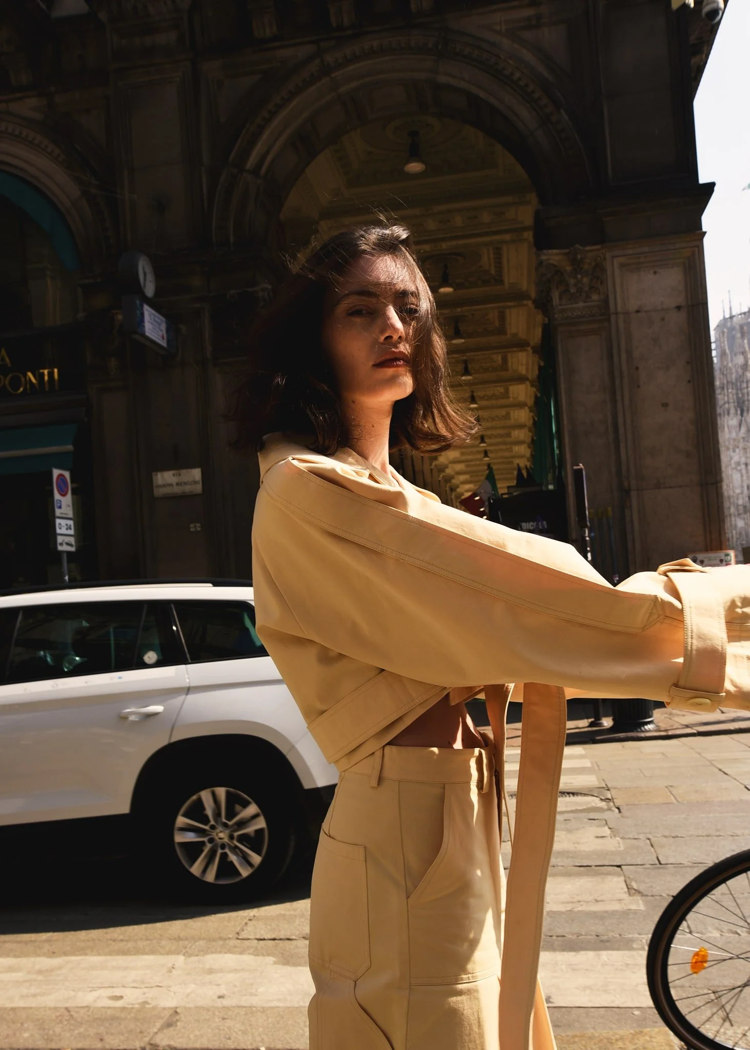 Donna in abbigliamento beige sulla strada con architettura storica sullo sfondo, auto e bicicletta vicino.