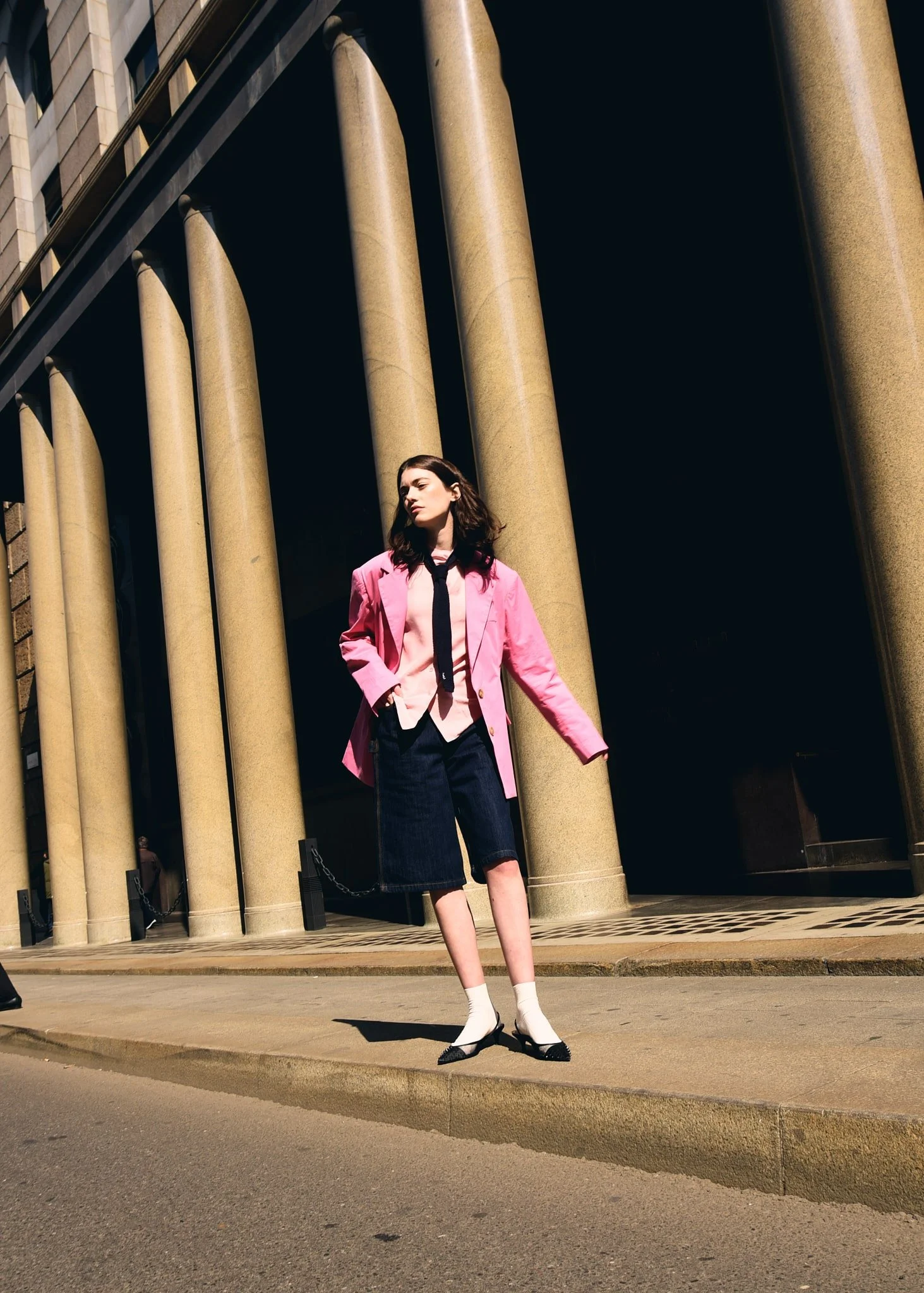 Una donna con indumenti eleganti, indecisa tra un abbigliamento formale e casual, si trova davanti a un edificio con grandi colonne in stile classico, sotto una luce solare intensa.