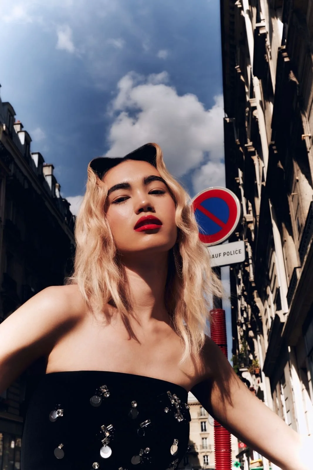 Una donna con capelli biondi e labbra rosse, indossa un vestito nero decorato con paillettes e un cappello a forma di gatto, in una strada urbana durante il giorno con un cielo nuvoloso.
