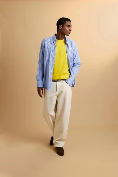 Uomo che indossa una camicia a righe blu sopra una maglietta gialla, pantaloni bianchi, e scarpe nere, che cammina di lato.