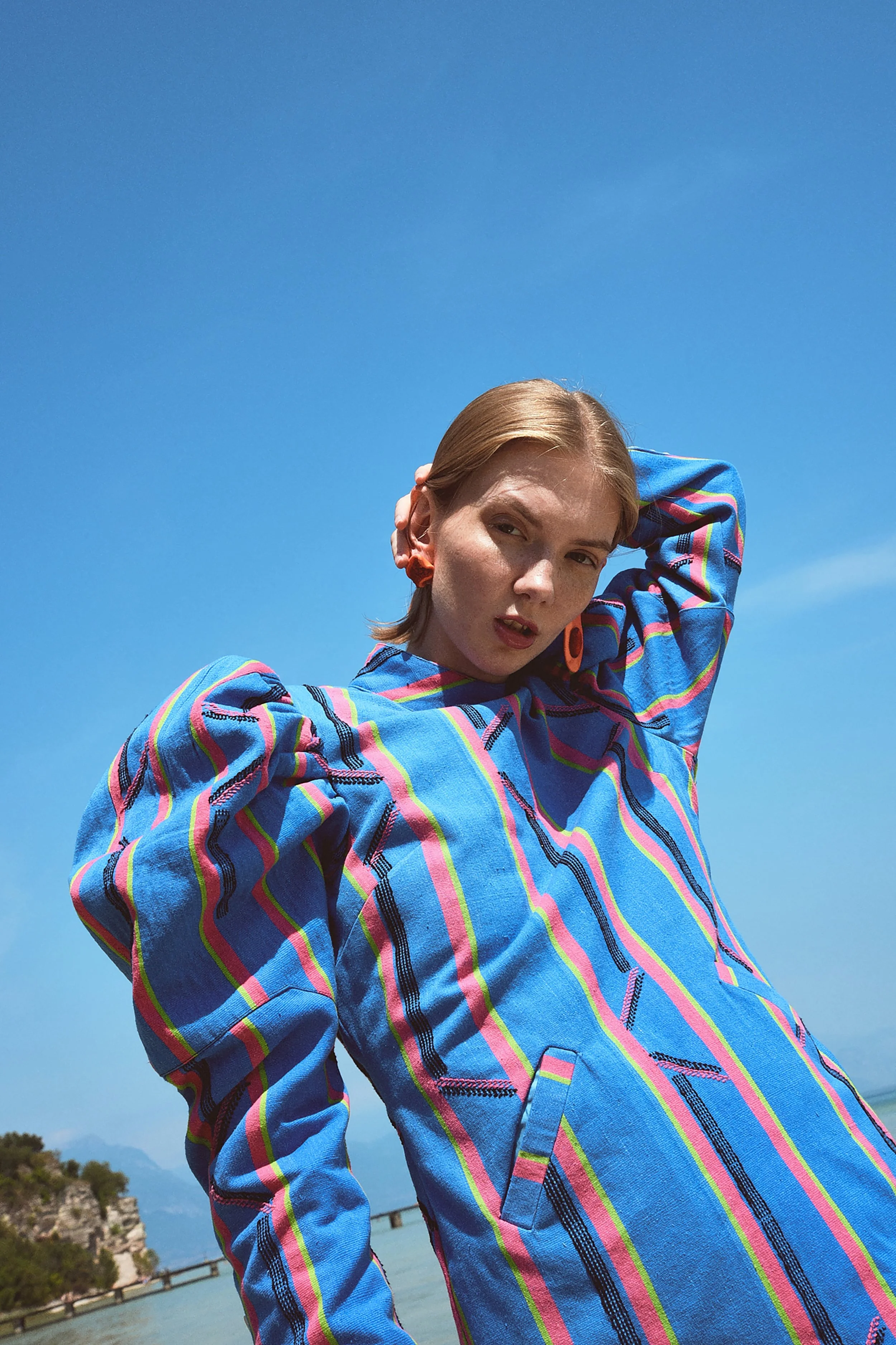 Ragazza con capelli corti e cappello rosso, indossa una giacca azzurra con motivi a righe colorate, sfondo di cielo azzurro e acqua.