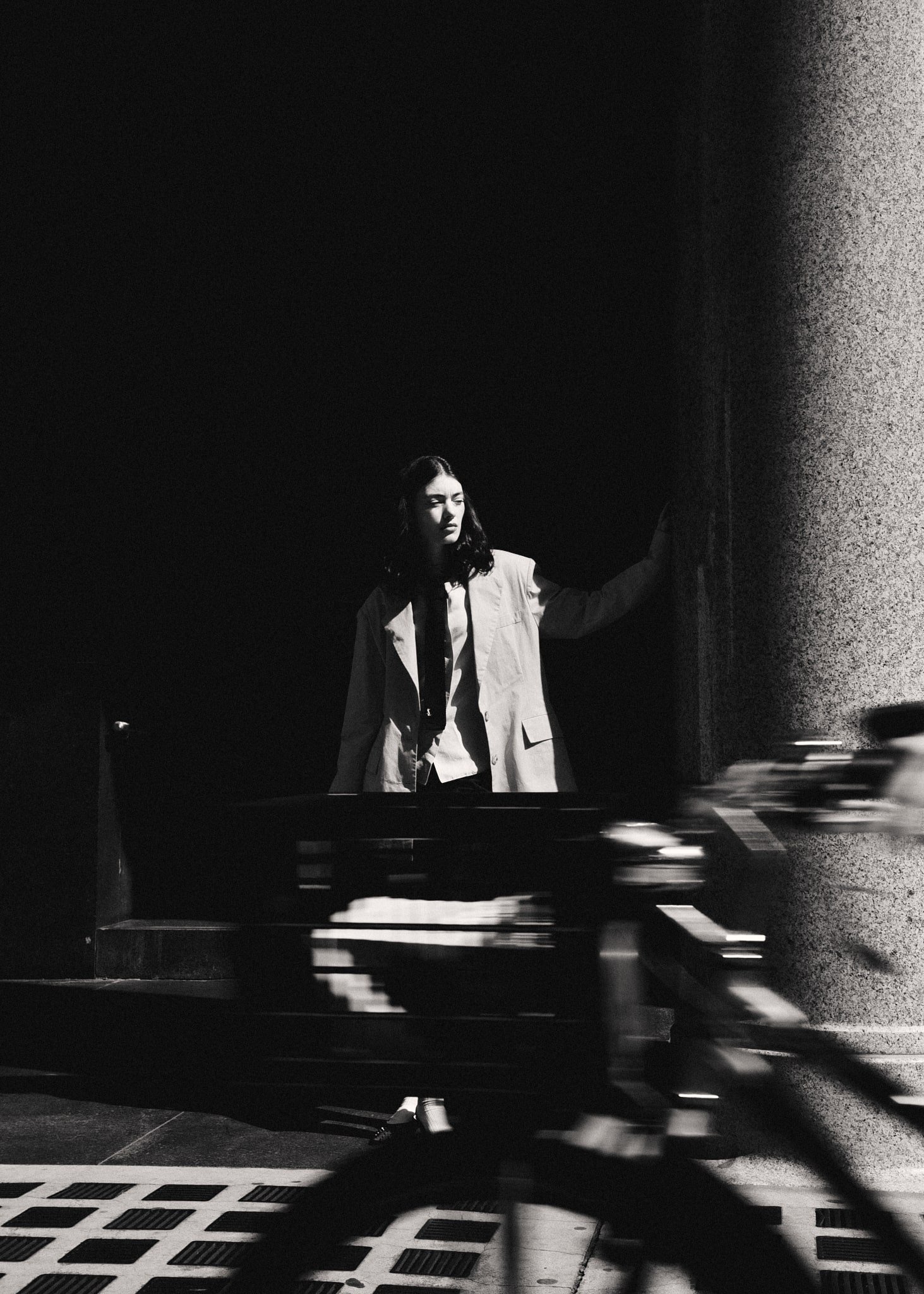 Donna in abito chiaro si appoggia a un muro in un'illuminazione drammatica in bianco e nero, con una bicicletta che passa di corsa davanti, creando un senso di movimento e contrasto.