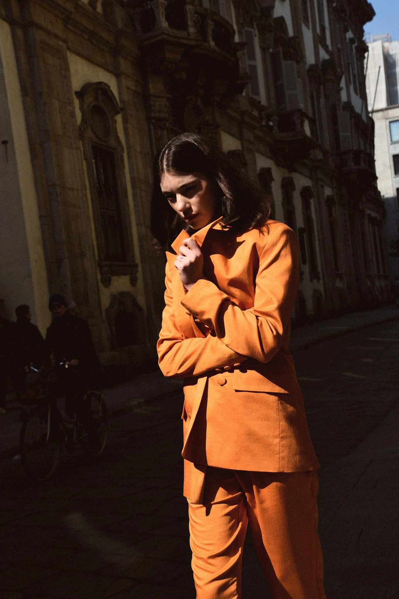Ragazza con capelli neri e pelle chiara indossa un completo arancione in una strada urbana con edifici storici sullo sfondo.