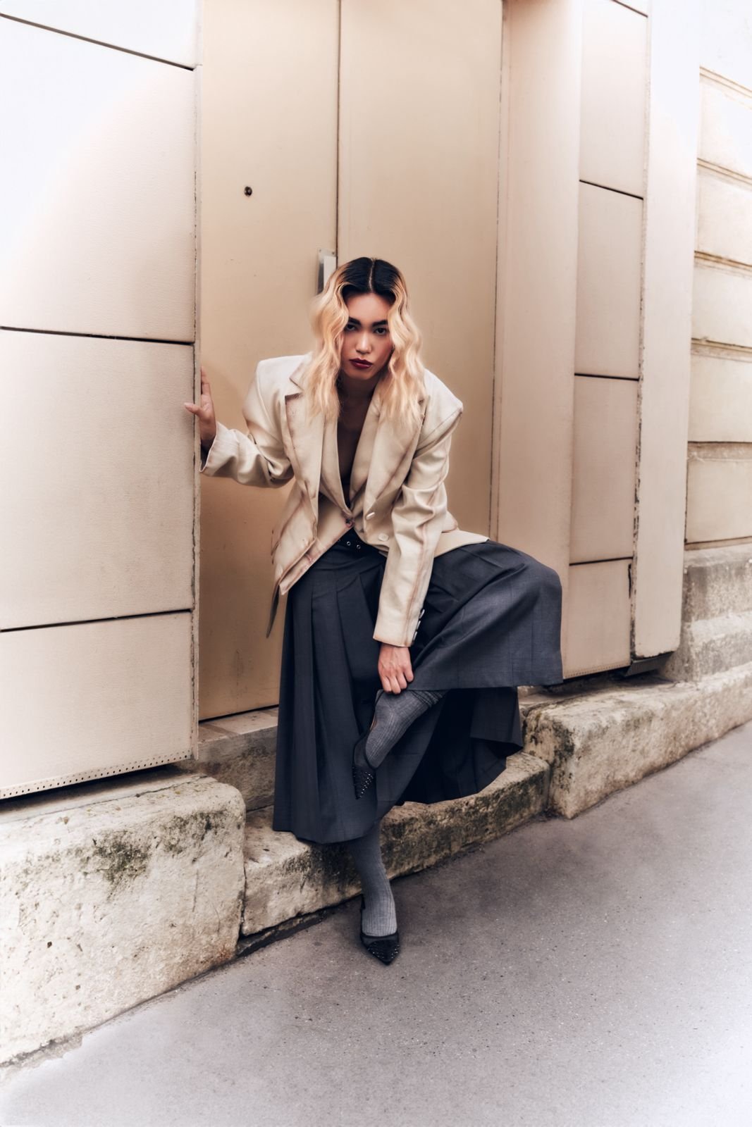 Donna seduta in abbigliamento formale con giacca beige, gonna lunga grigia, calze a righe e scarpe con tacco, contro un muro beige in città.