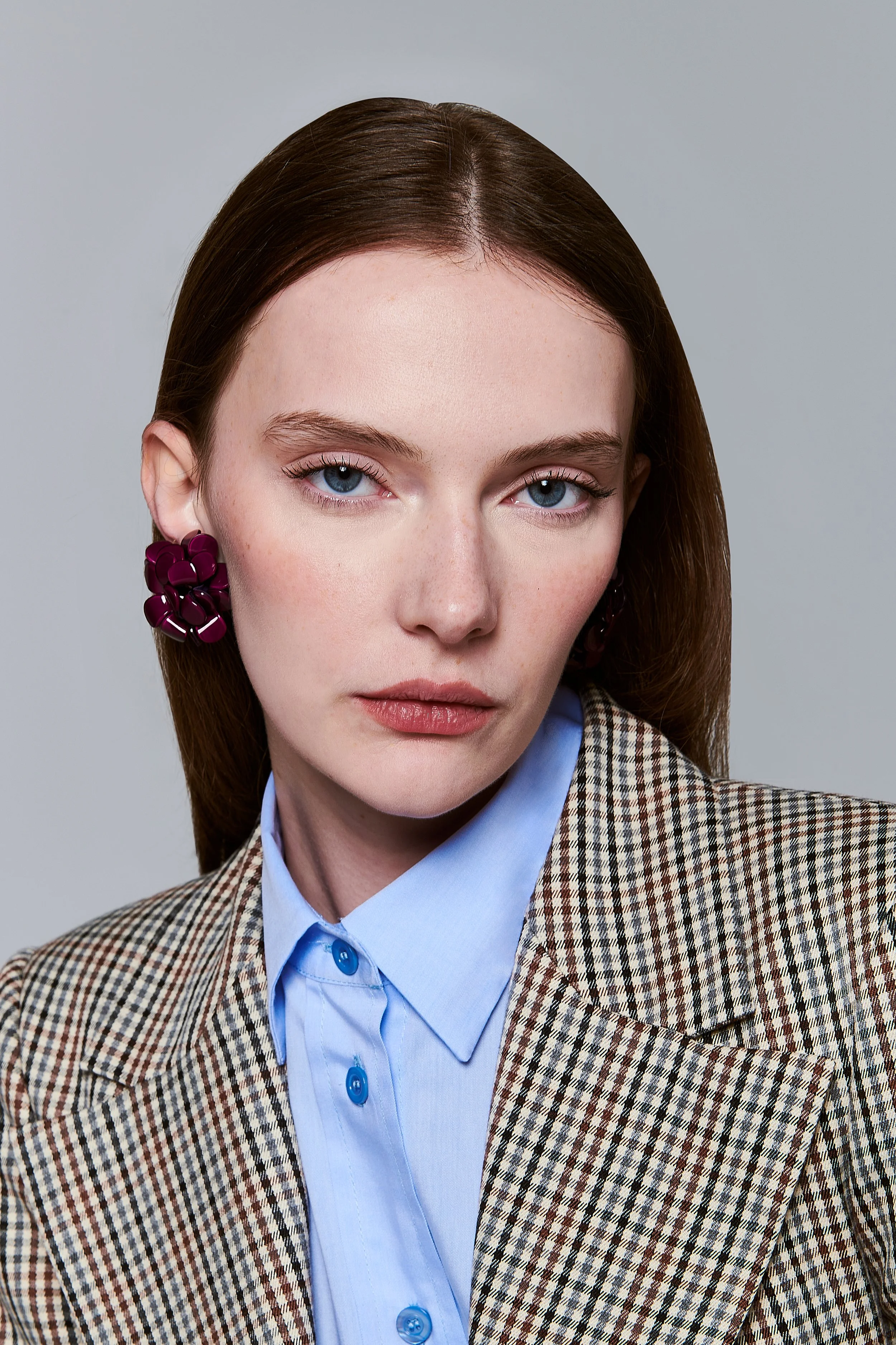 Ragazza con capelli castani e occhi azzurri, indossa orecchini viola a forma di fiore, camicia azzurra e giacca a quadretti.