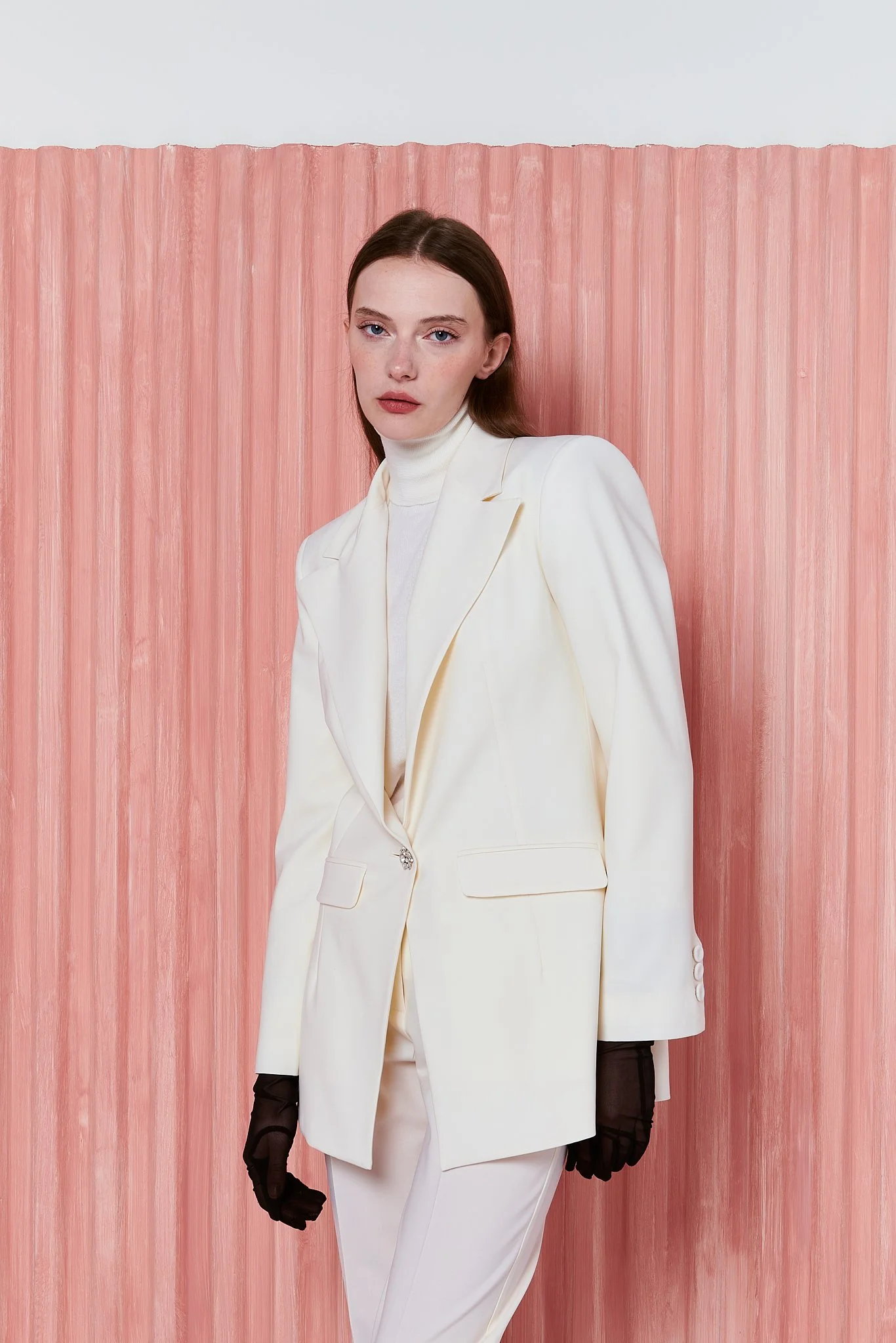 Donna con abito bianco, blazer e pantaloni, con guanti neri, posando di fronte a uno sfondo rosa stratificato.