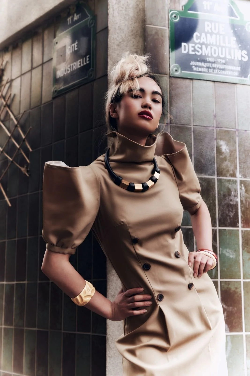 Una donna giovane con capelli biondi raccolti in un impeto disordinato, indossa un vestito beige con maniche a puffy e dettagli in nero, con accessori come collana nera e bracciali colorati, posa con una mano sulla cinta su uno sfondo di pareti di ma