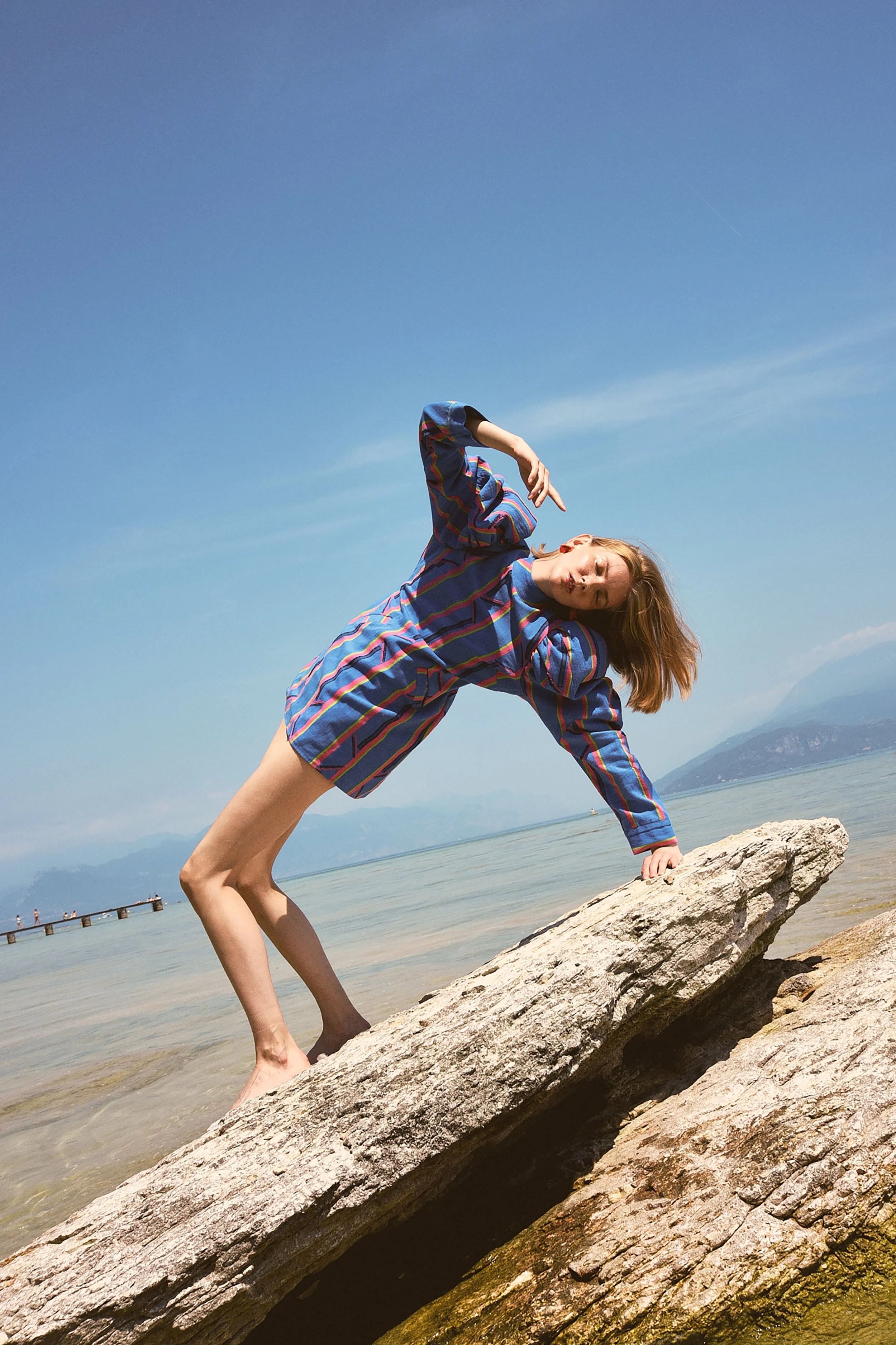 Una donna con un abito a righe blu, rossi e verdi si appoggia su una roccia vicina al lago sotto un cielo sereno.