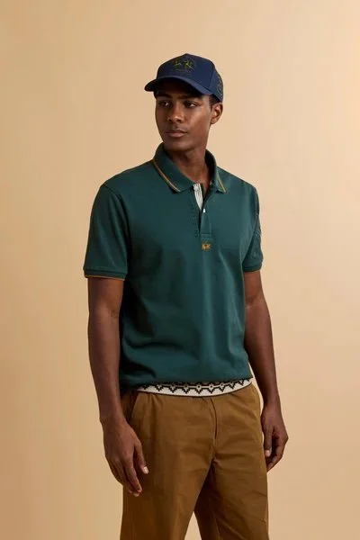 Un uomo giovane vestito con polo verde, cappellino blu e pantaloni marroni posa contro uno sfondo beige.