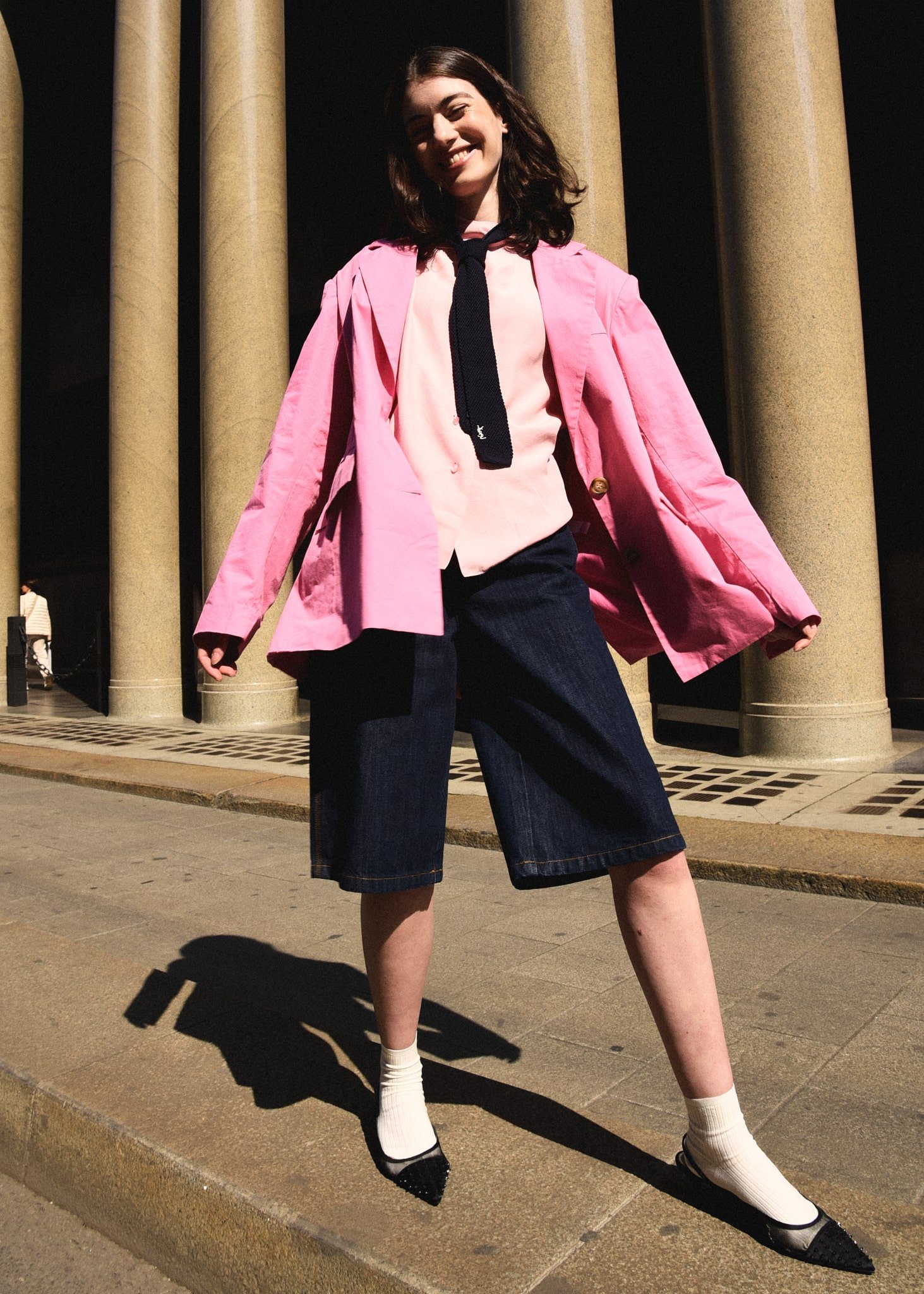 Una donna sorridente con un cappotto rosa, camicia rosa, cravatta nera, gonna lunga di jeans e scarpe con calze bianche, si trova davanti a colonne di pietra di notte.