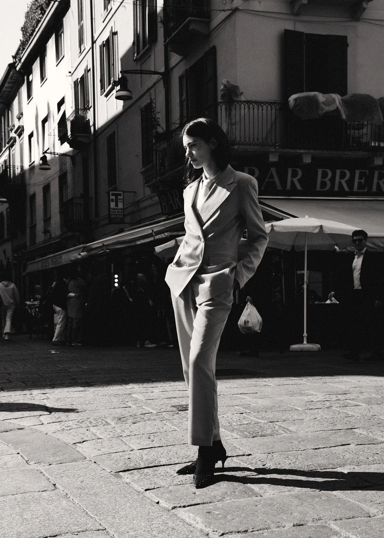 Donna vestita in abiti eleganti camminando per strada in una città, con edifici storici sullo sfondo, in una foto in bianco e nero.