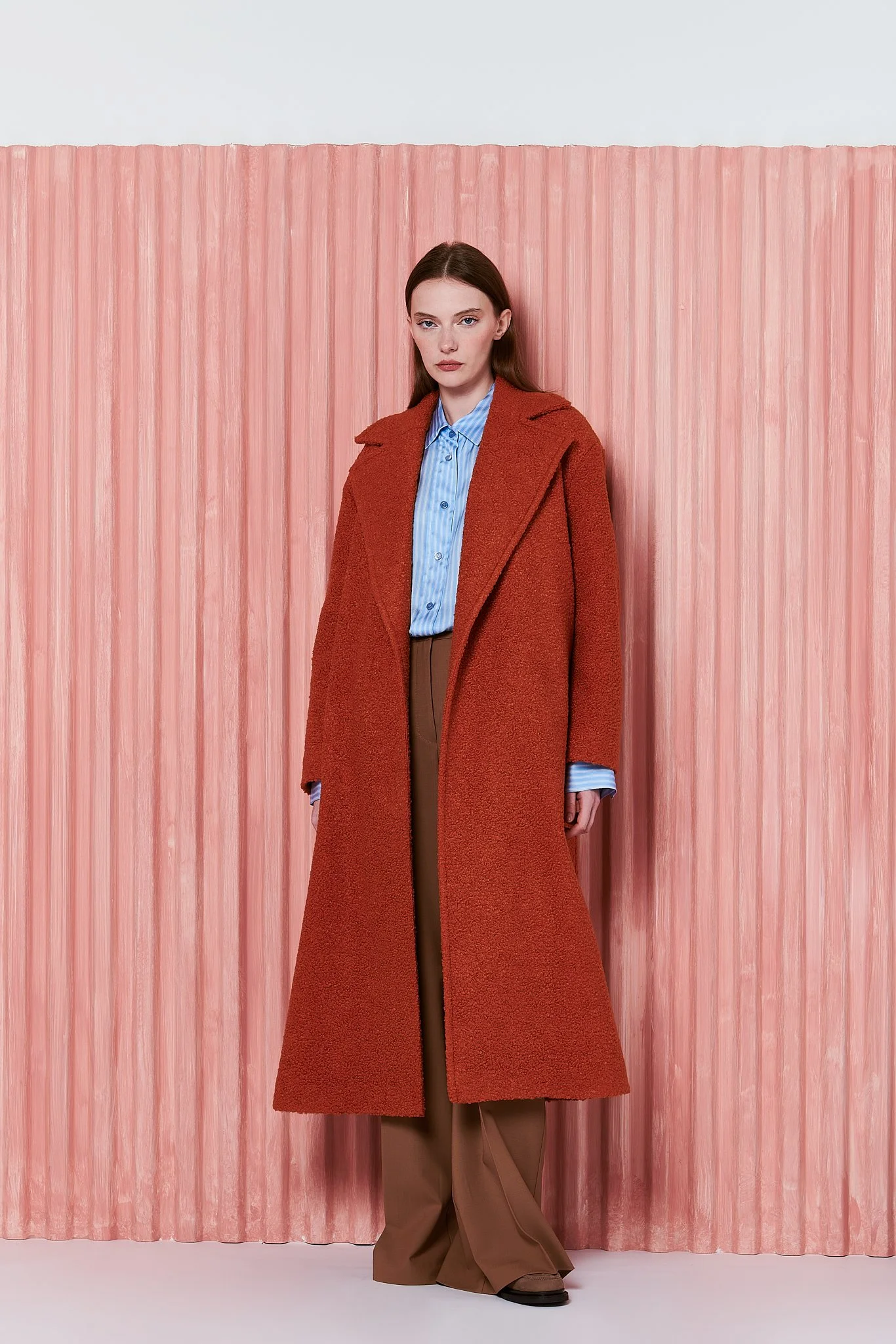 Donna con calzamaglia a vita alta, camicia azzurra a righe, e cappotto rosso lungo in piedi davanti a una parete rosa