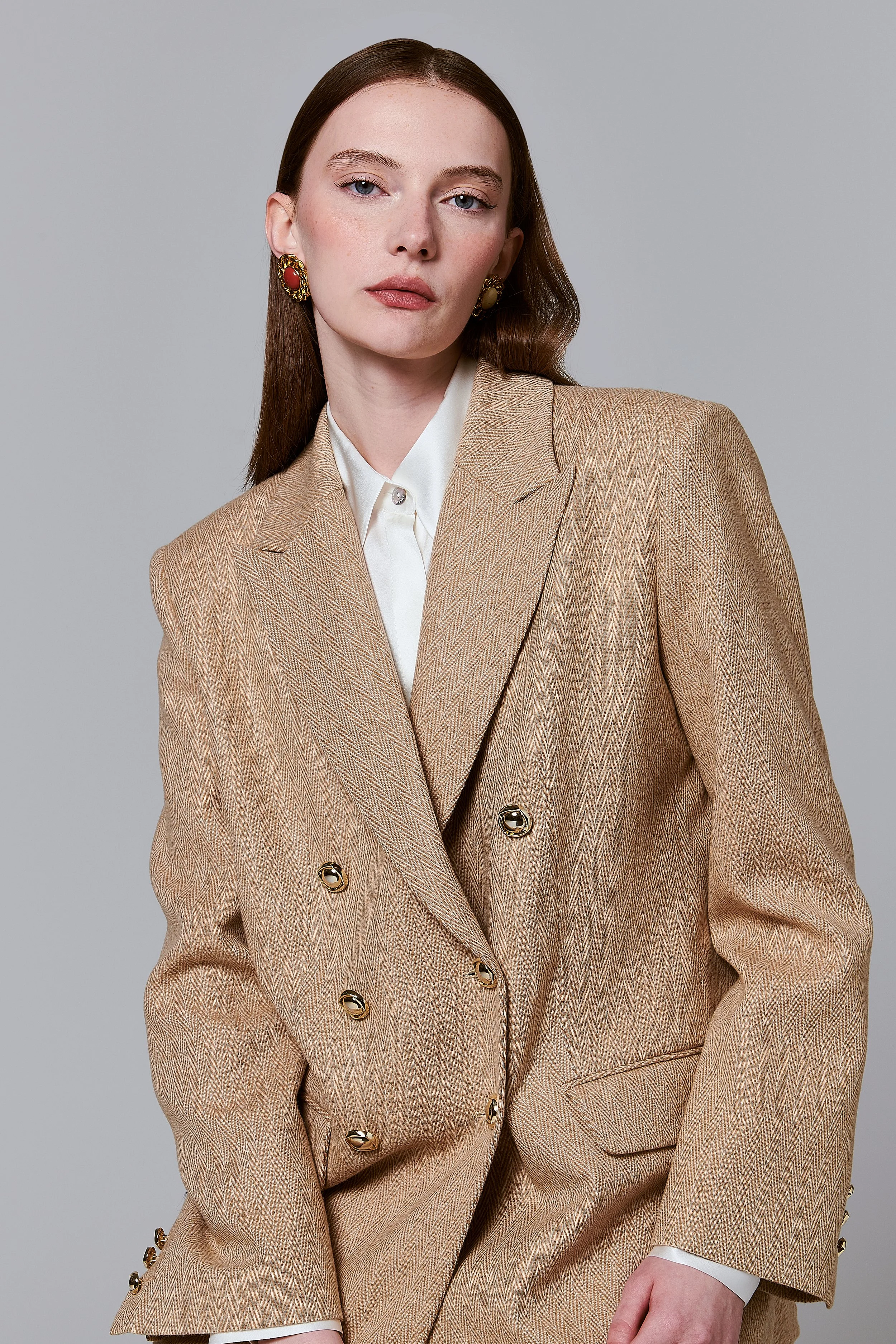 Donna con capelli castani lunghi, indossa un completo beige con giacca doppiopetto e camicia bianca, anche con orecchini dorati e rossi.