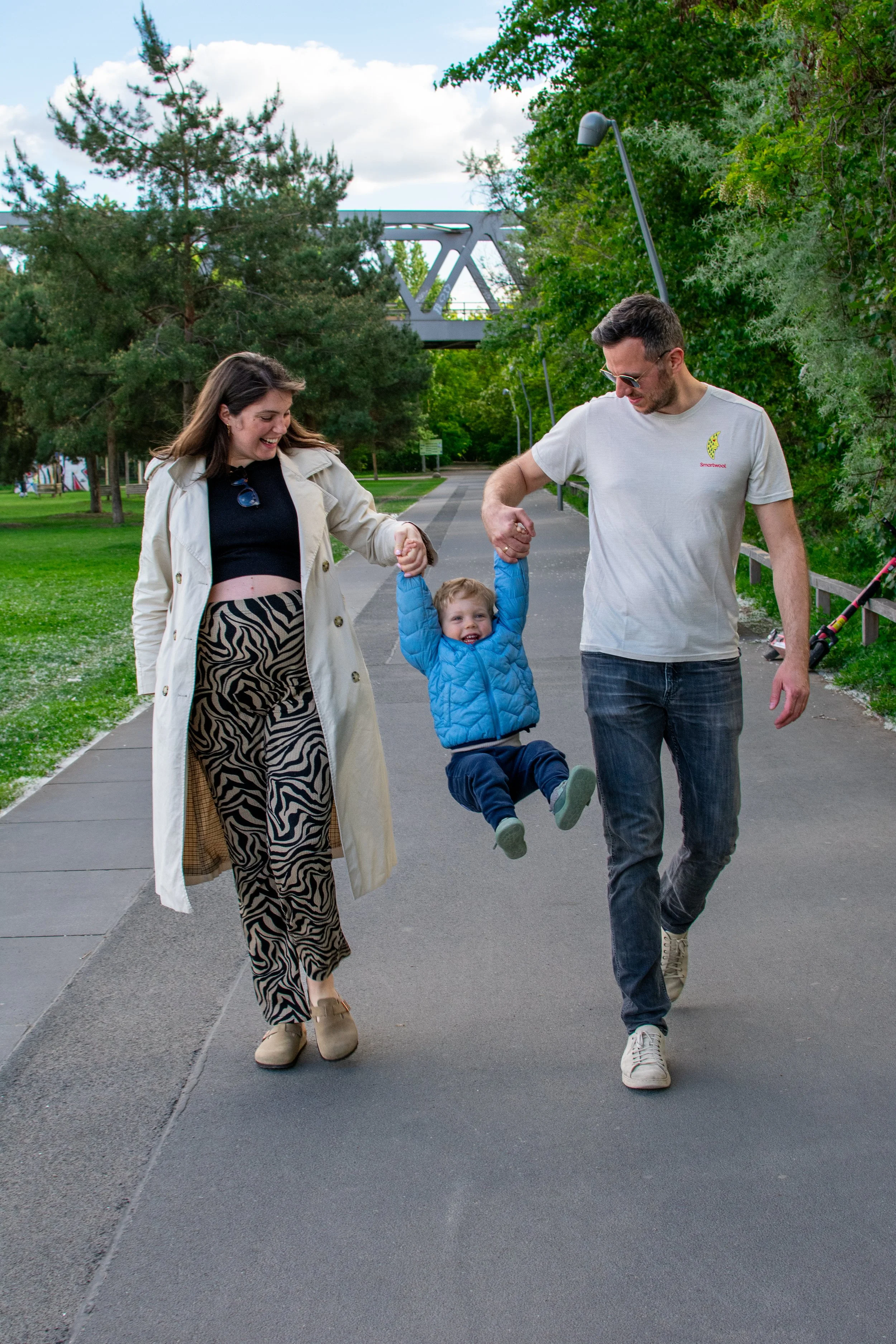 A family of three on a park walkway, with a woman and a man holding a young child by the hands as the child swings between them, smiling joyfully.