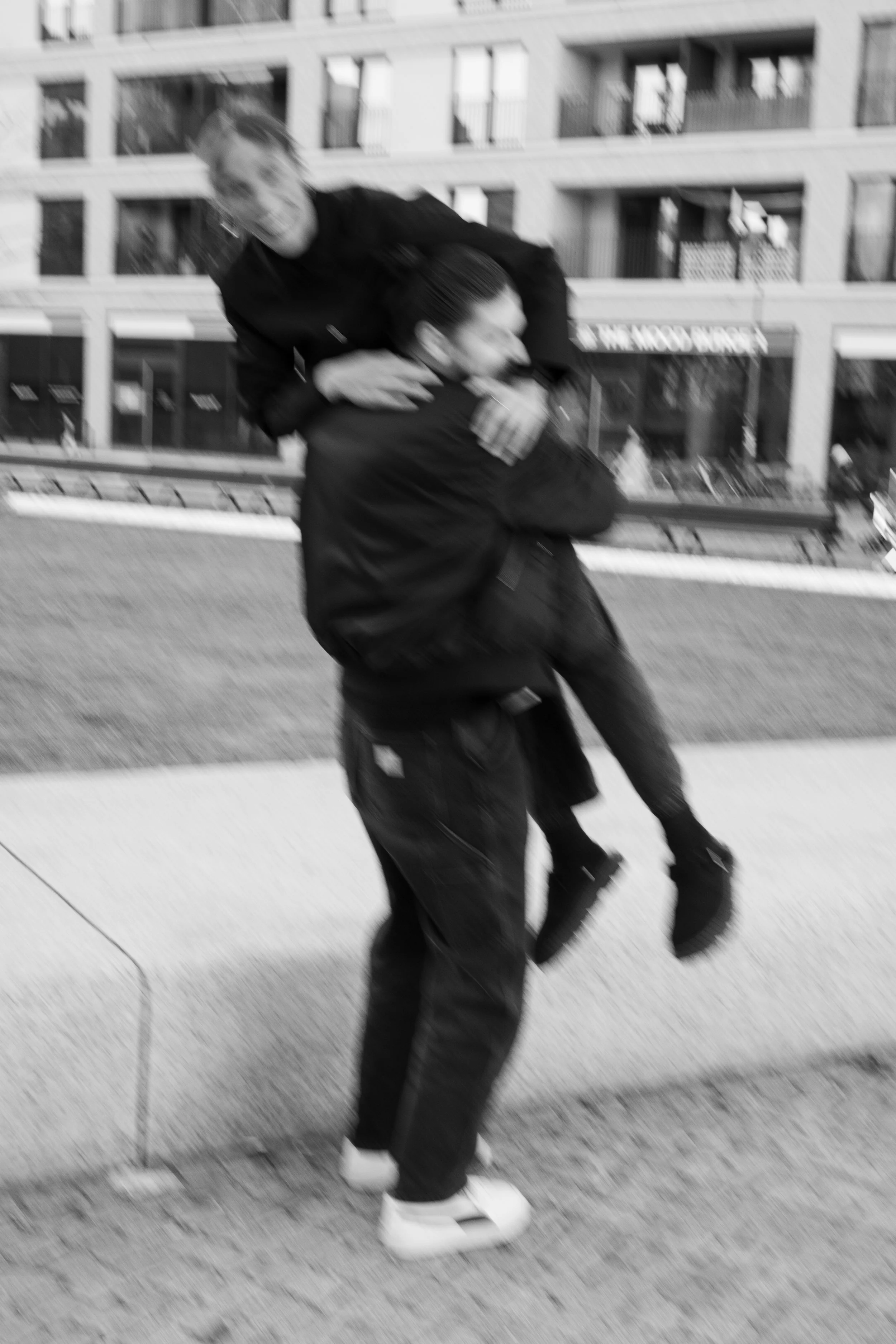 Two people, one lifting the other, on a city sidewalk with multi-story buildings in the background