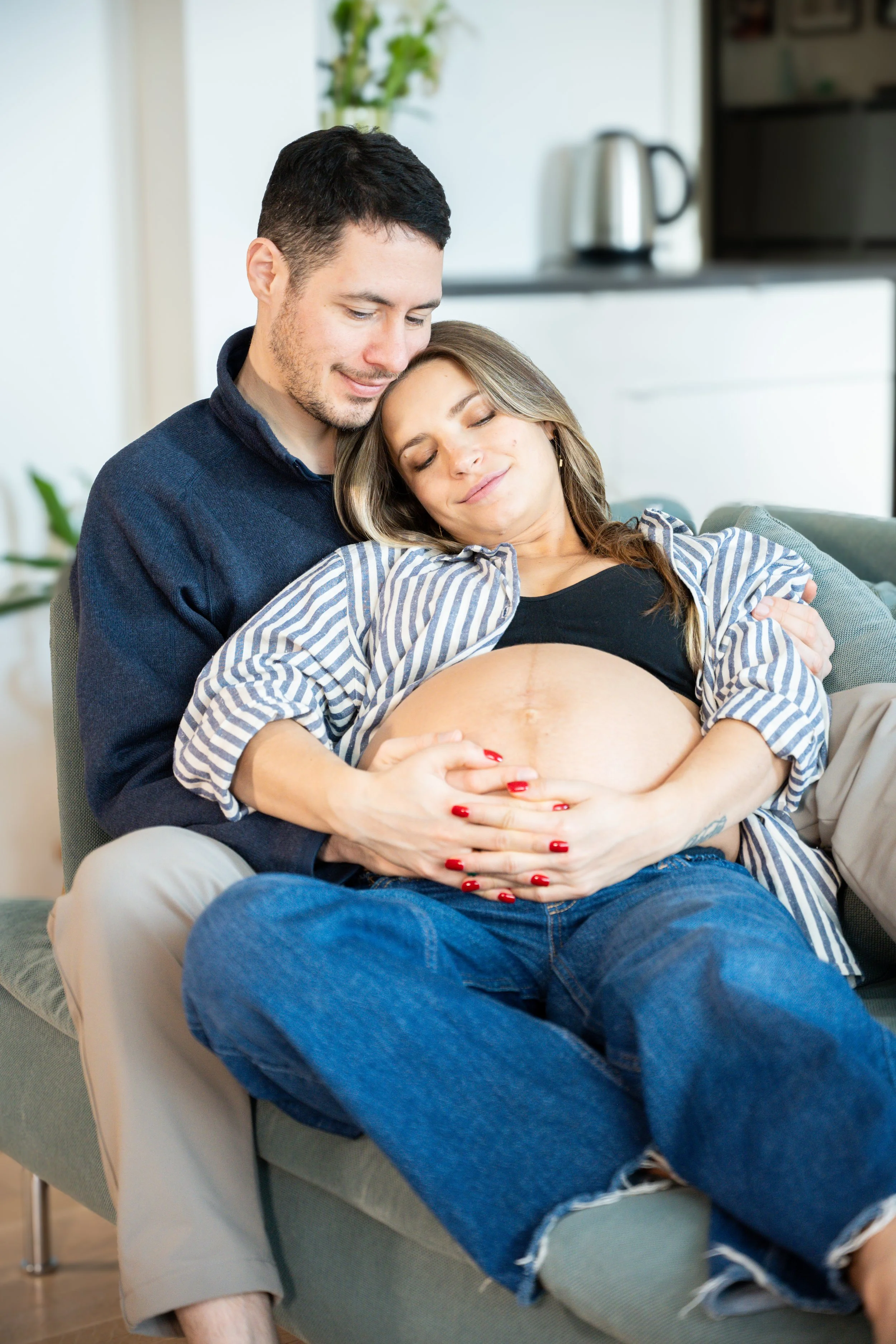 A pregnant woman with closed eyes and a content smile sits on a couch, cradling her belly, while a man sitting behind her gently embraces her and places his hands on her belly. Both are smiling softly in a cozy home setting.