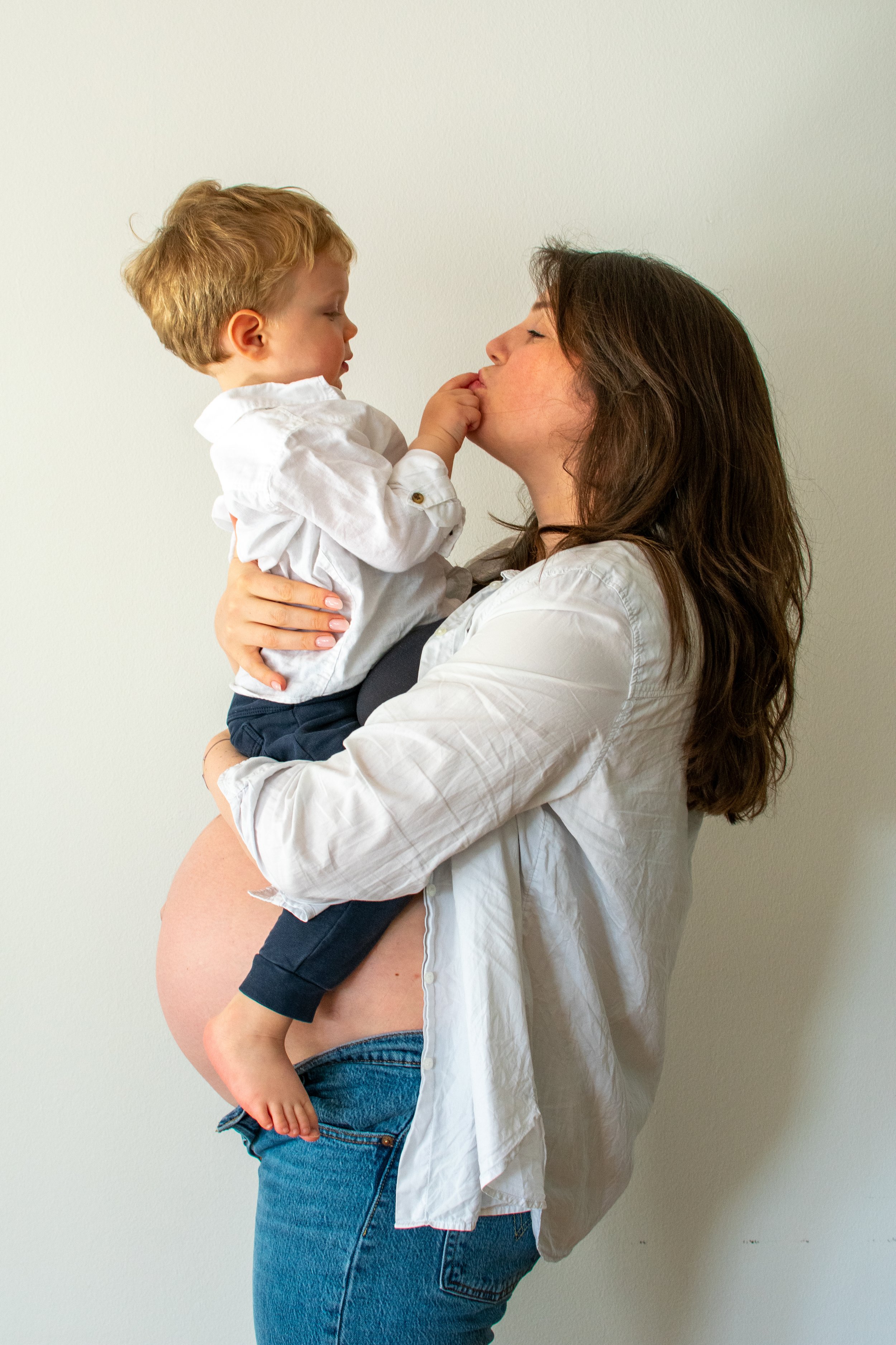 A smiling woman holding a young boy, who is touching her nose, against a plain light-colored wall.