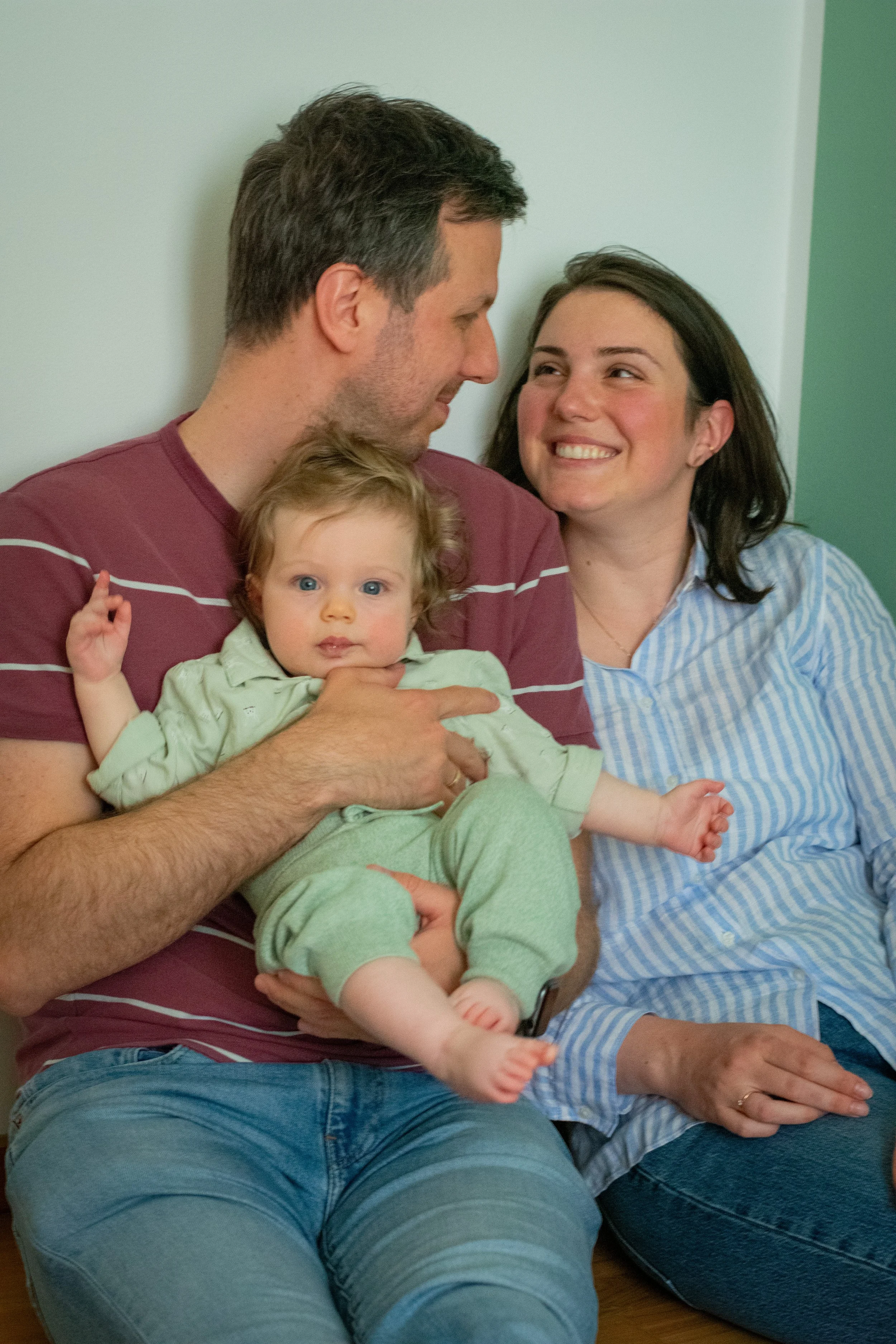 A man, a woman, and a baby sitting together indoors. The man is holding the baby, and all three are smiling and looking at each other. The woman is sitting next to them, smiling warmly.