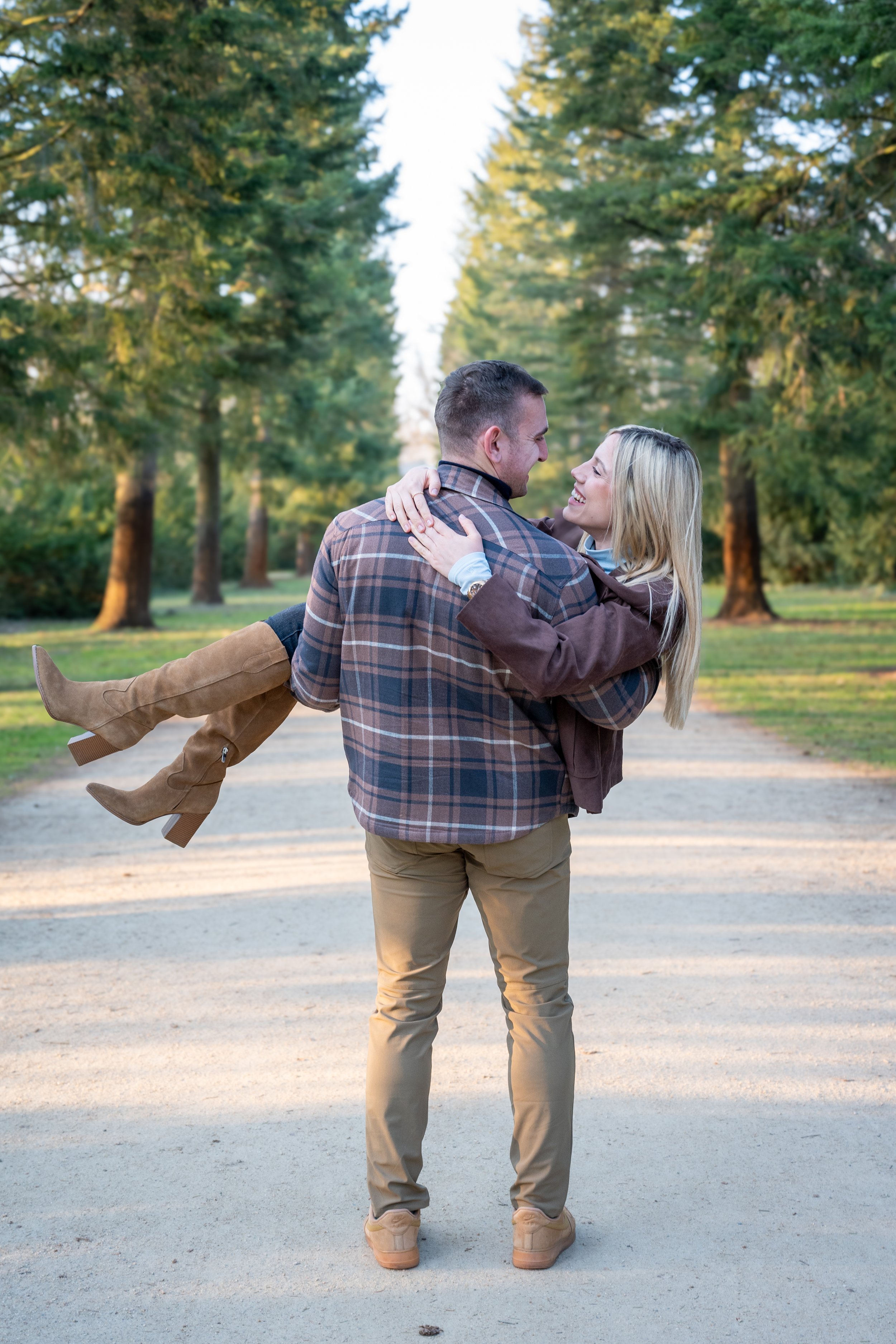 A happy couple in a park, with the man carrying the woman as they smile at each other surrounded by green trees.