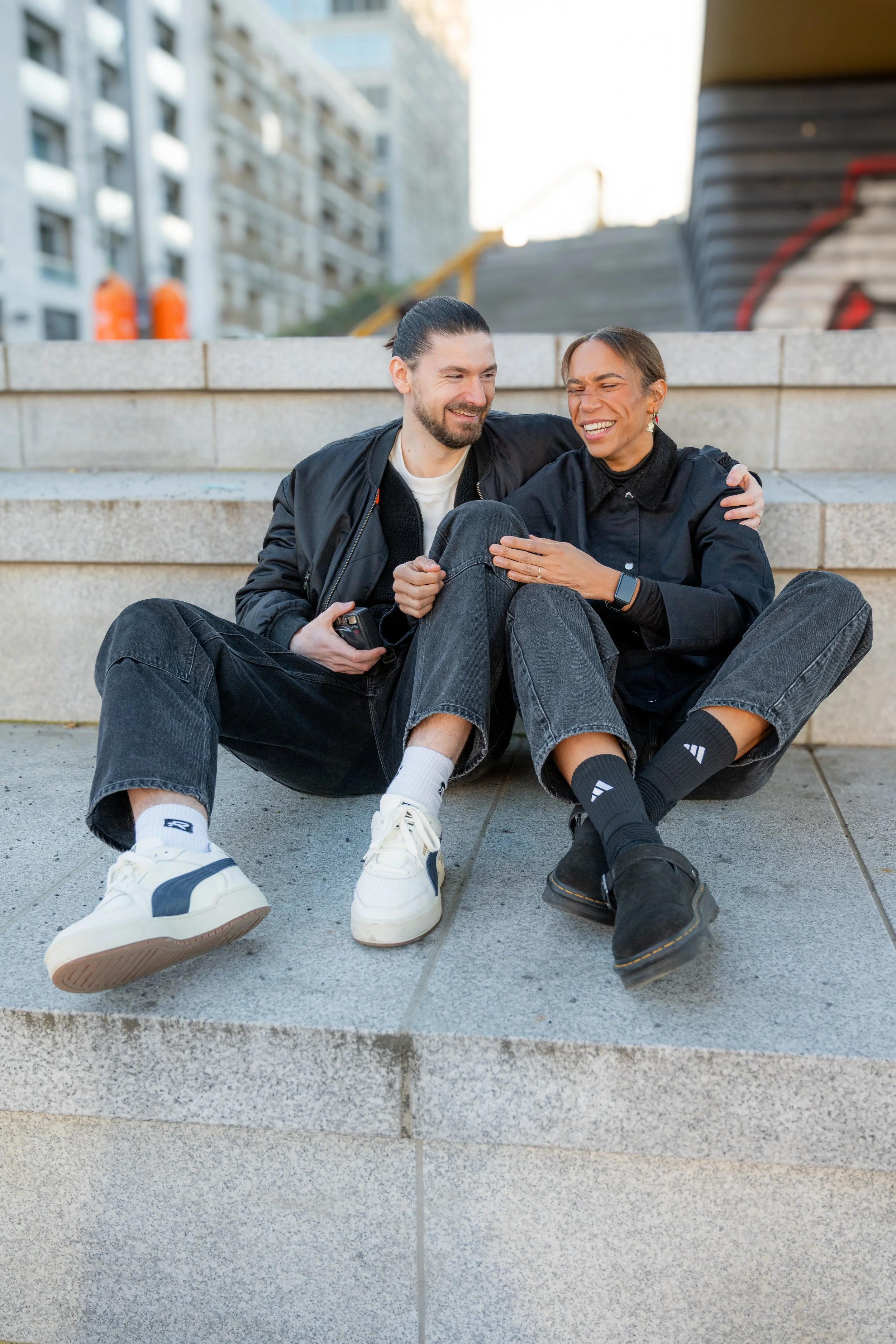 A man and woman sitting on concrete steps outdoors, smiling and laughing together, with city buildings in the background.