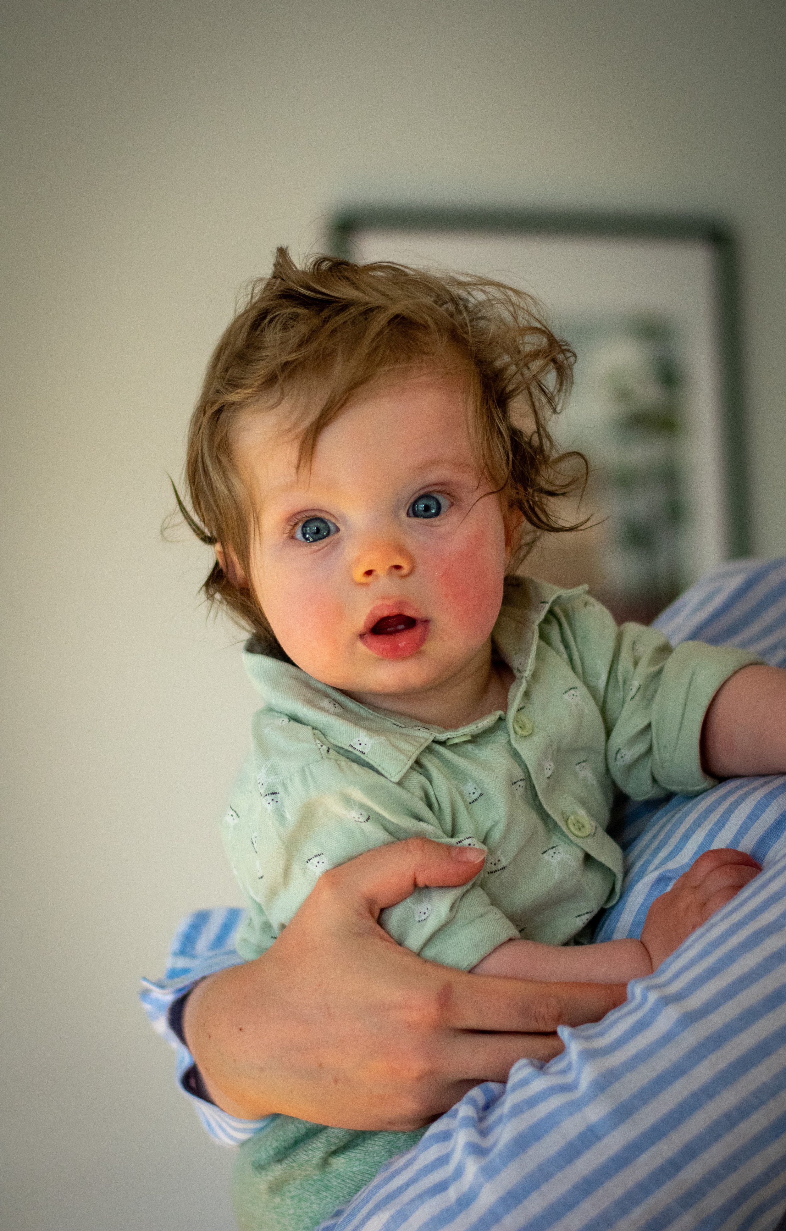 A young child with curly hair and blue eyes looks surprised or curious while being held by an adult, with a blurred indoor background.