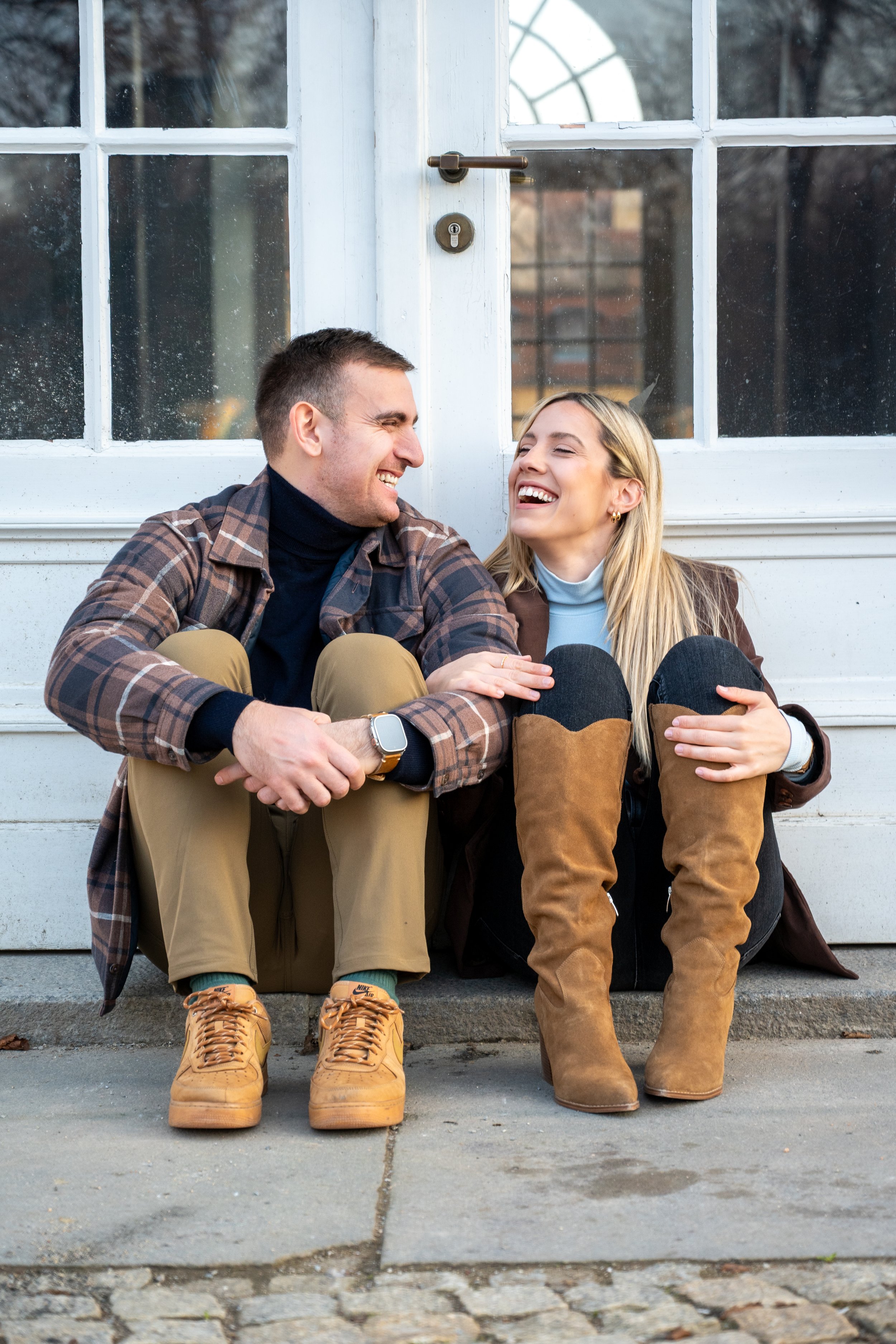 A young man and woman sitting on the sidewalk in front of a white door, sharing a joyful moment and laughing together.
