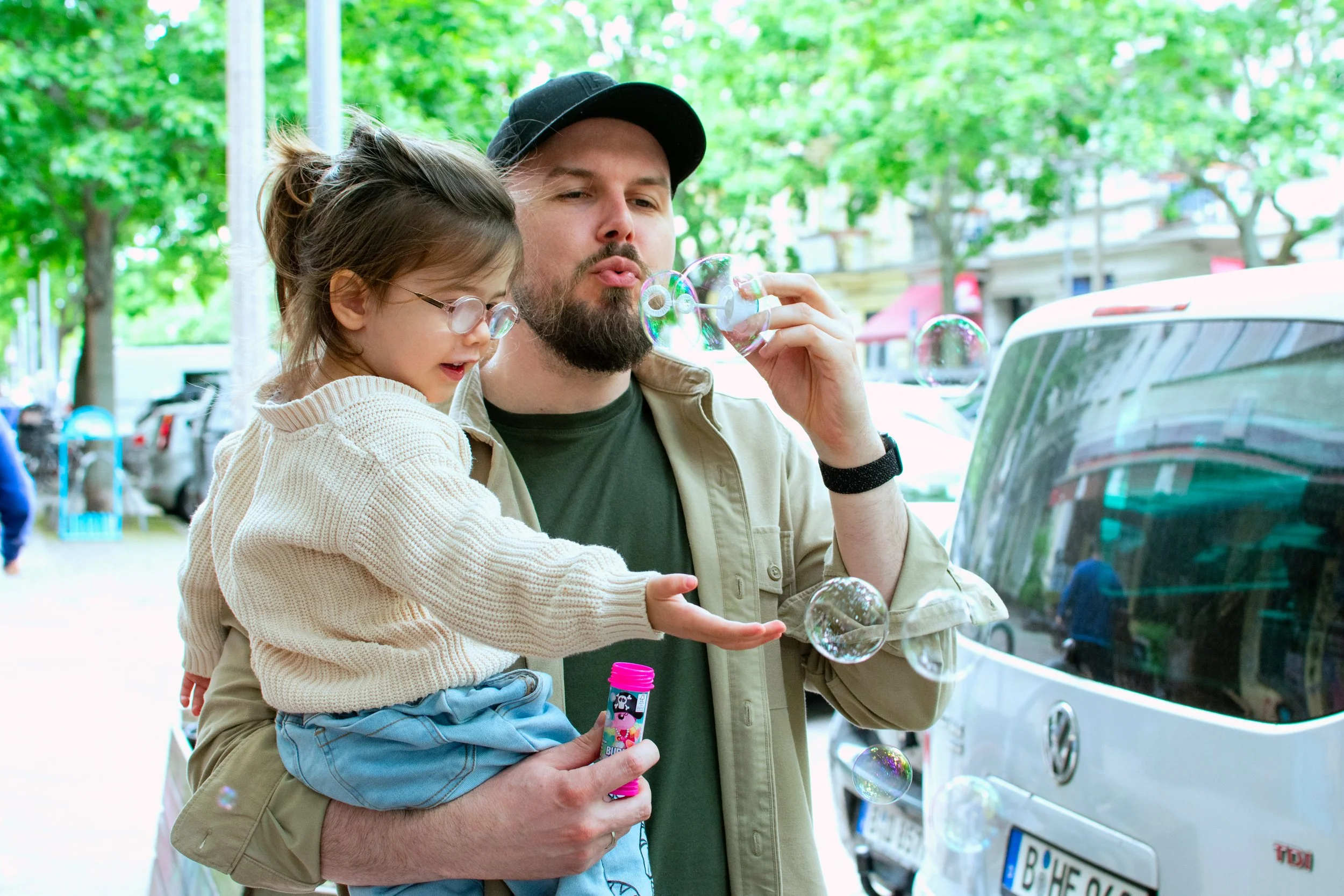 A man with a beard wearing a beige jacket and black cap holds a young girl in a cream sweater and glasses, both blowing bubbles outdoors near a white vehicle and green trees.