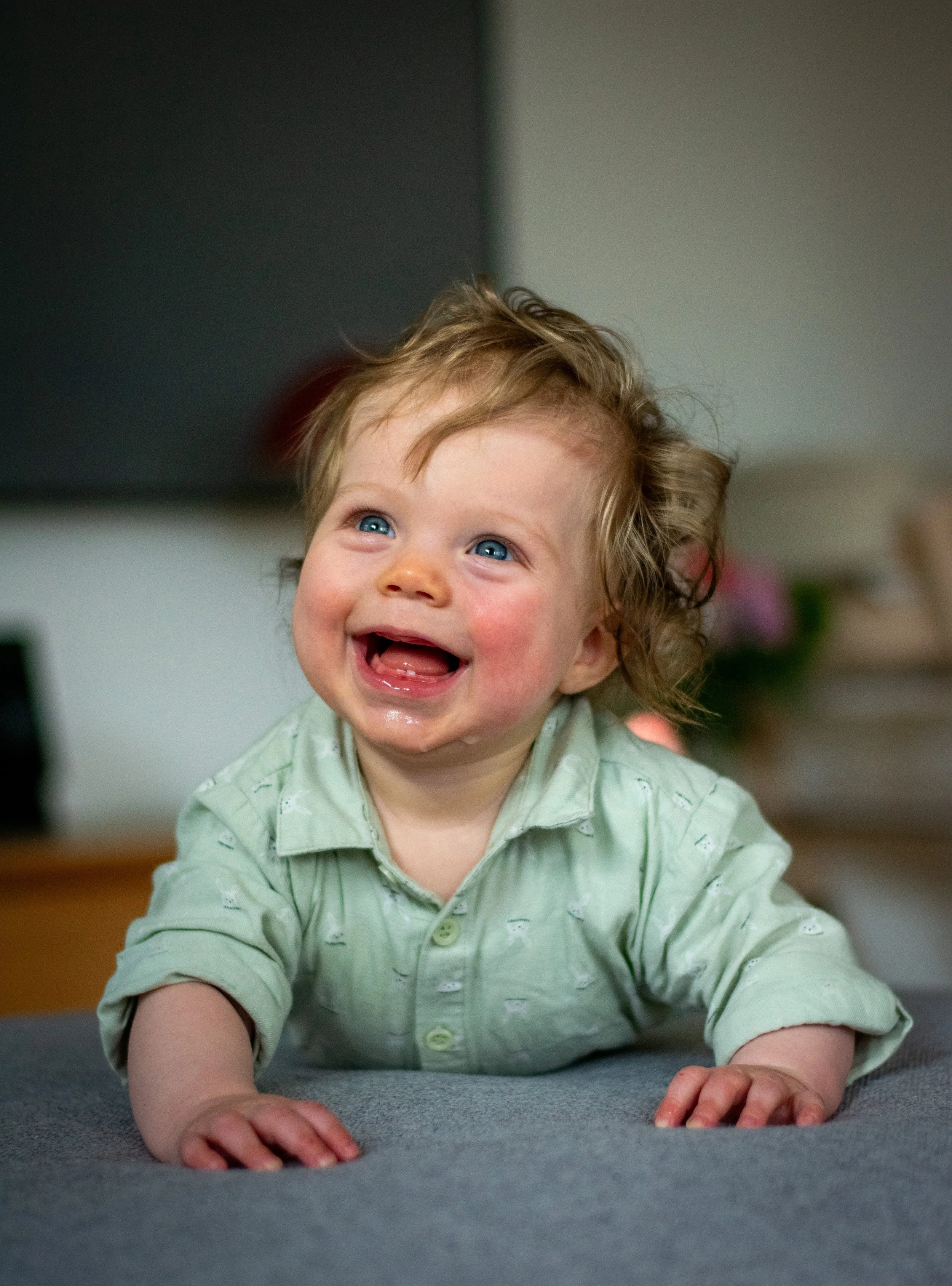 A happy baby with blue eyes, curly blond hair, wearing a light green shirt, laying on a gray surface indoors.