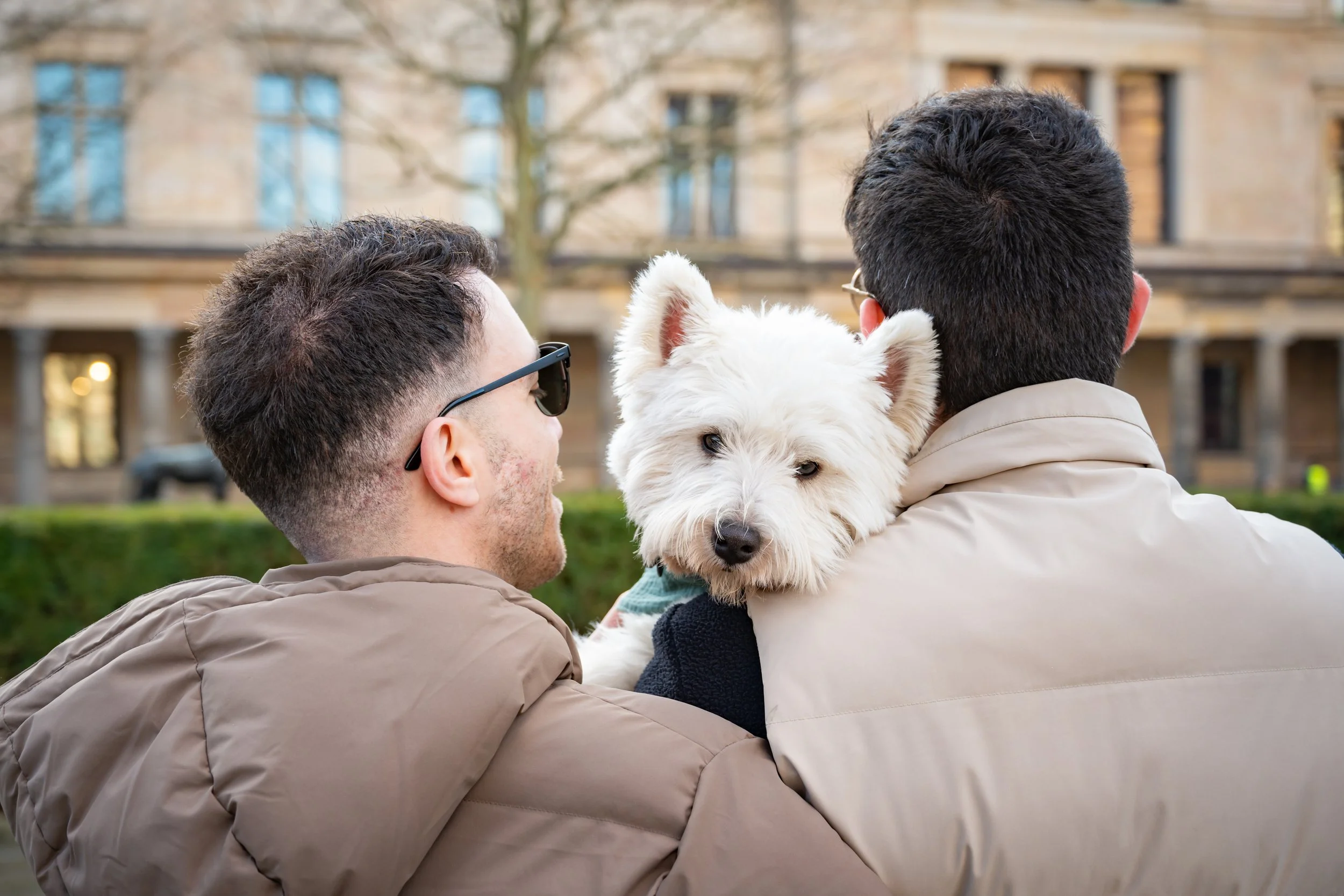 Two men holding a white dog with a coat outdoors in front of a building with trees and parked cars.
