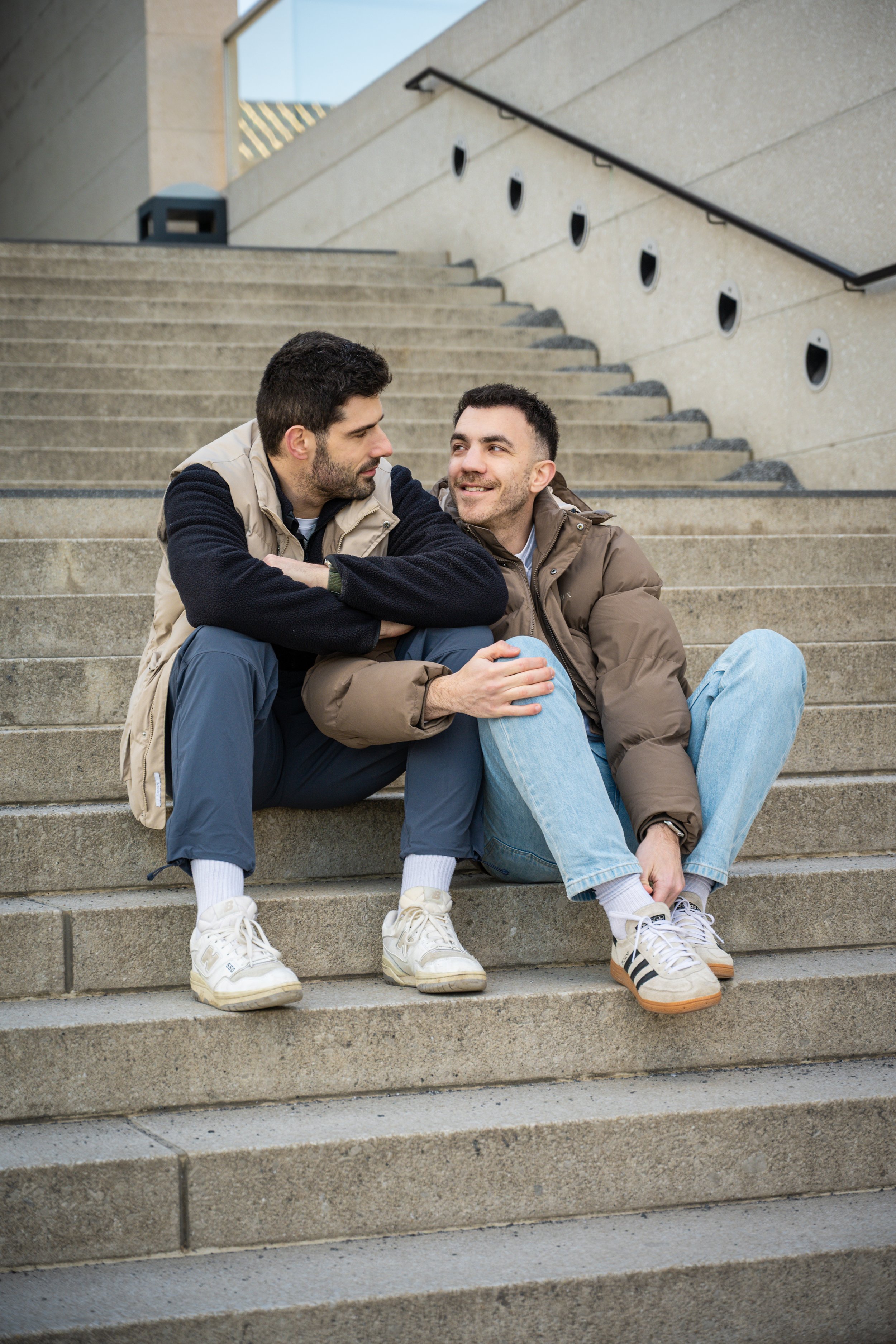 Two men sitting on outdoor concrete stairs, conversing and smiling at each other in casual clothing.