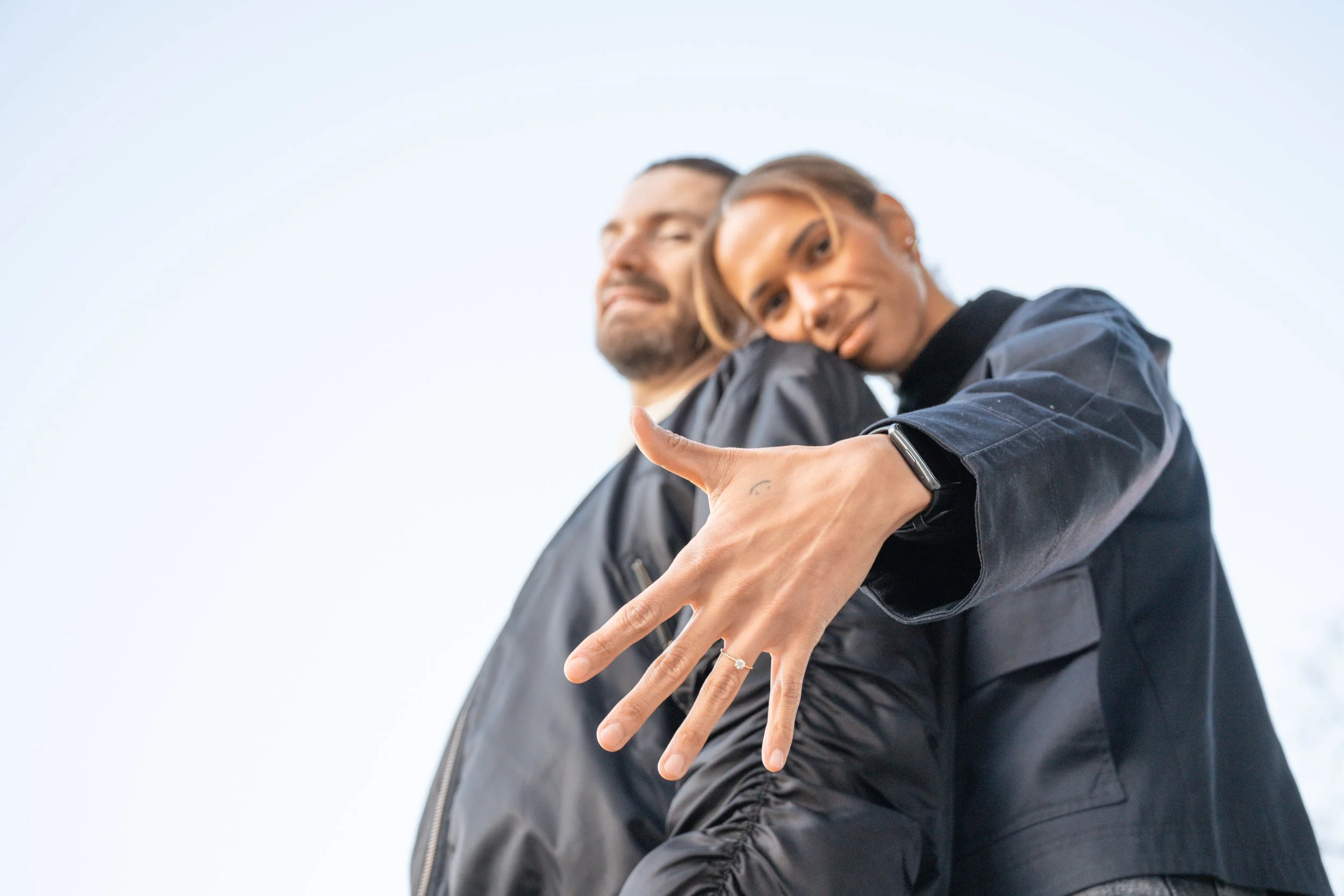 A woman with a watch and ring showing her hand in focus, making a peace sign, with a man in the background smiling, both wearing black jackets, against a clear sky.