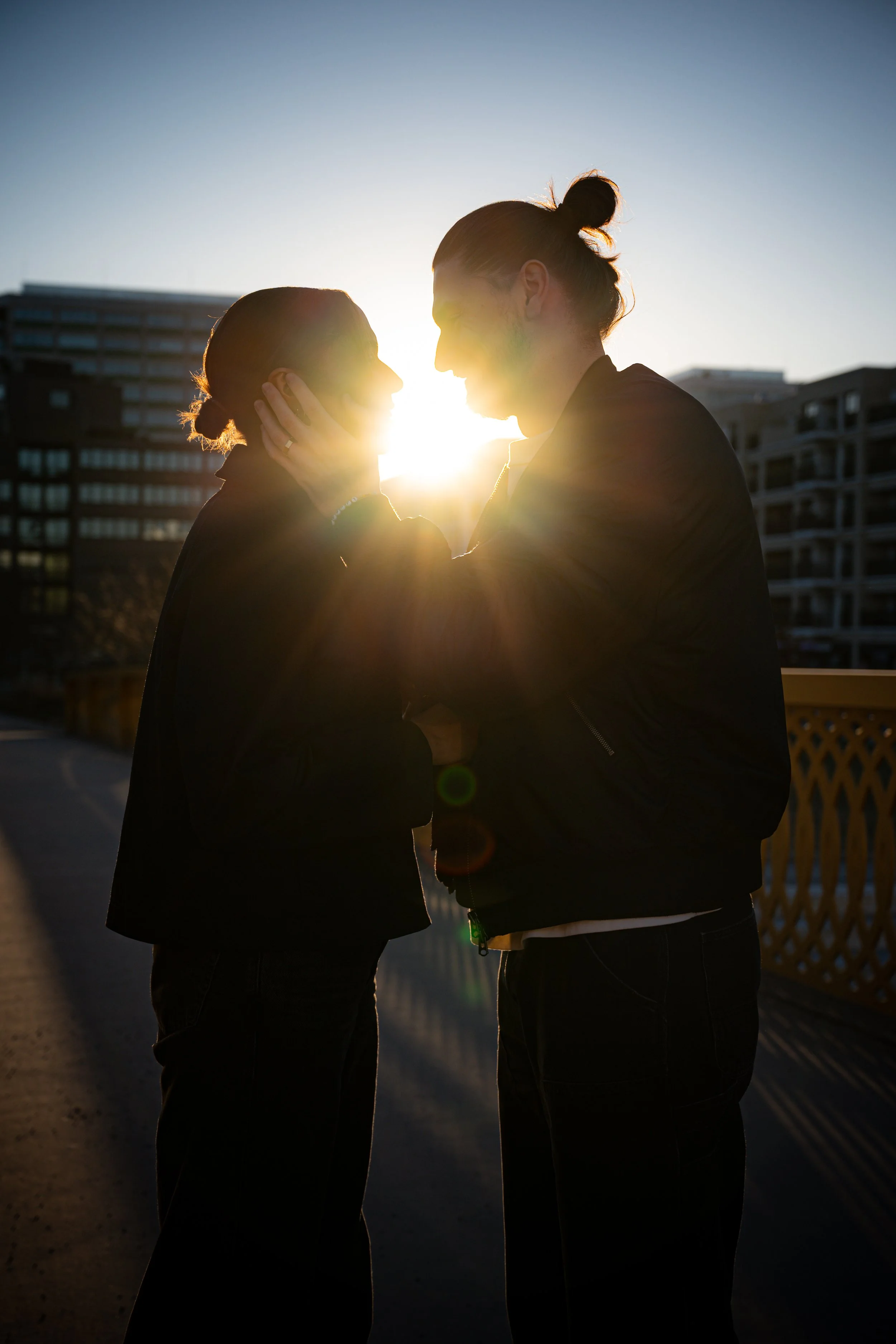 A couple during sunset, facing each other closely, with one holding the other's face, against an urban background.