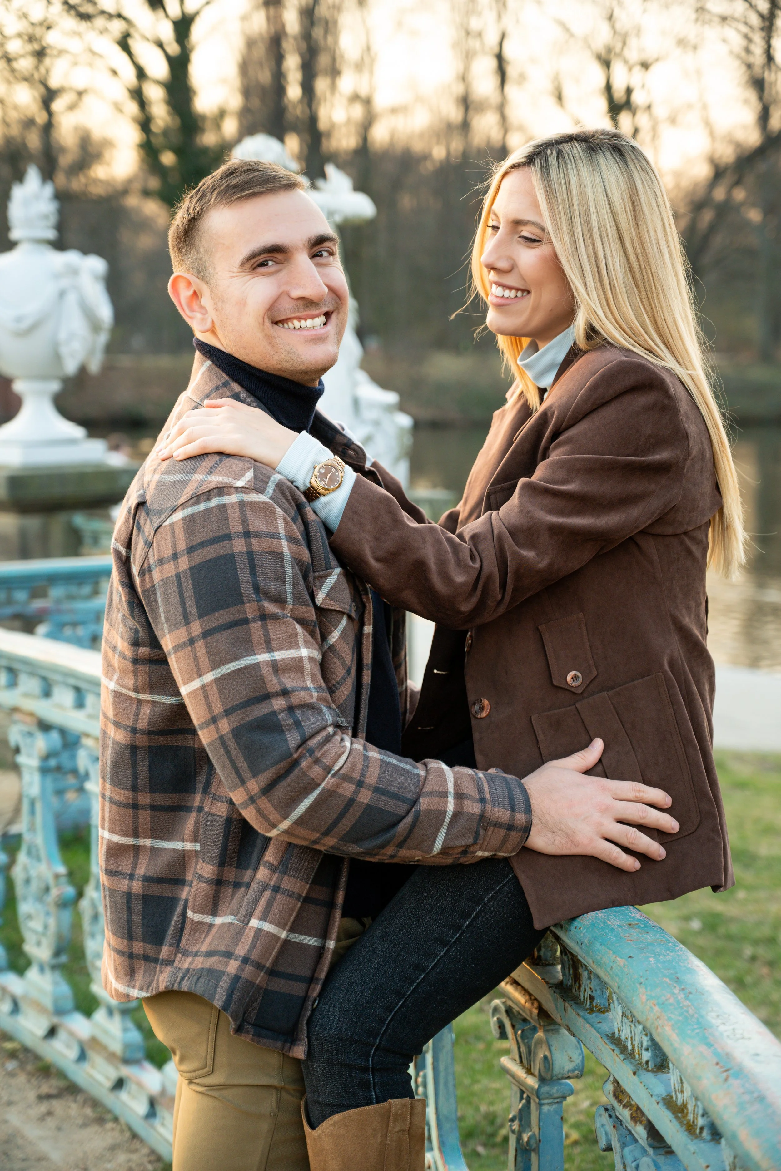 A happy couple smiling and embracing outdoors near a decorative railing and lake, during sunset.
