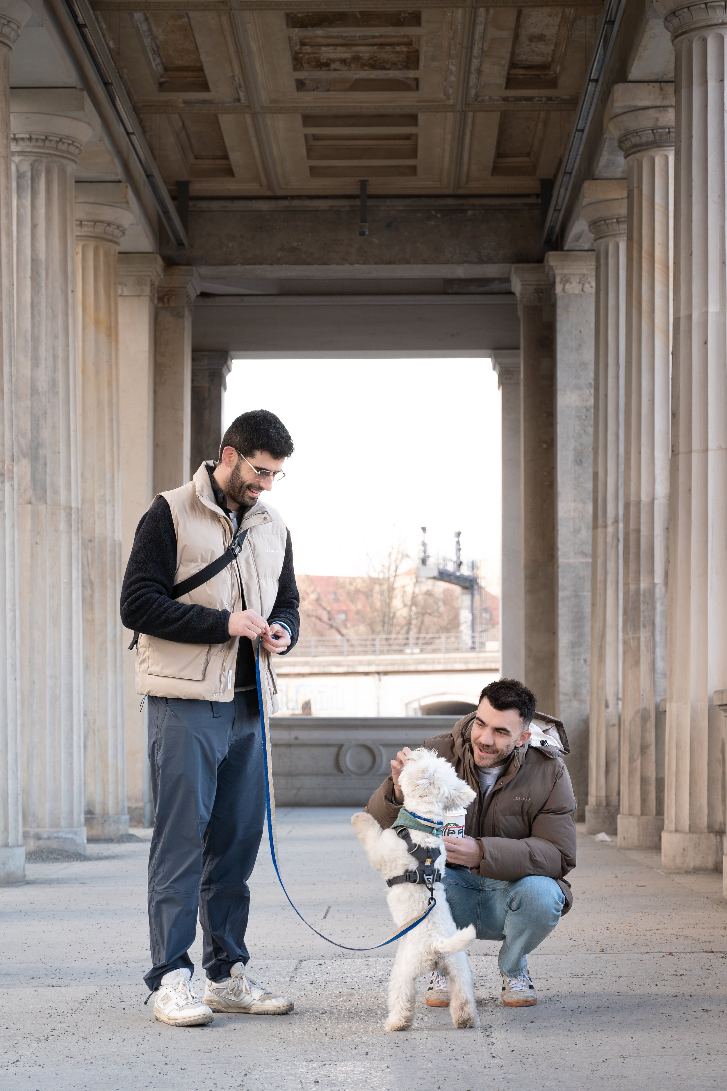 Two men with a white dog in a classical stone building with columns. One man is standing and holding the dog's leash, the other man is crouching and petting the dog. The dog is wearing a harness.