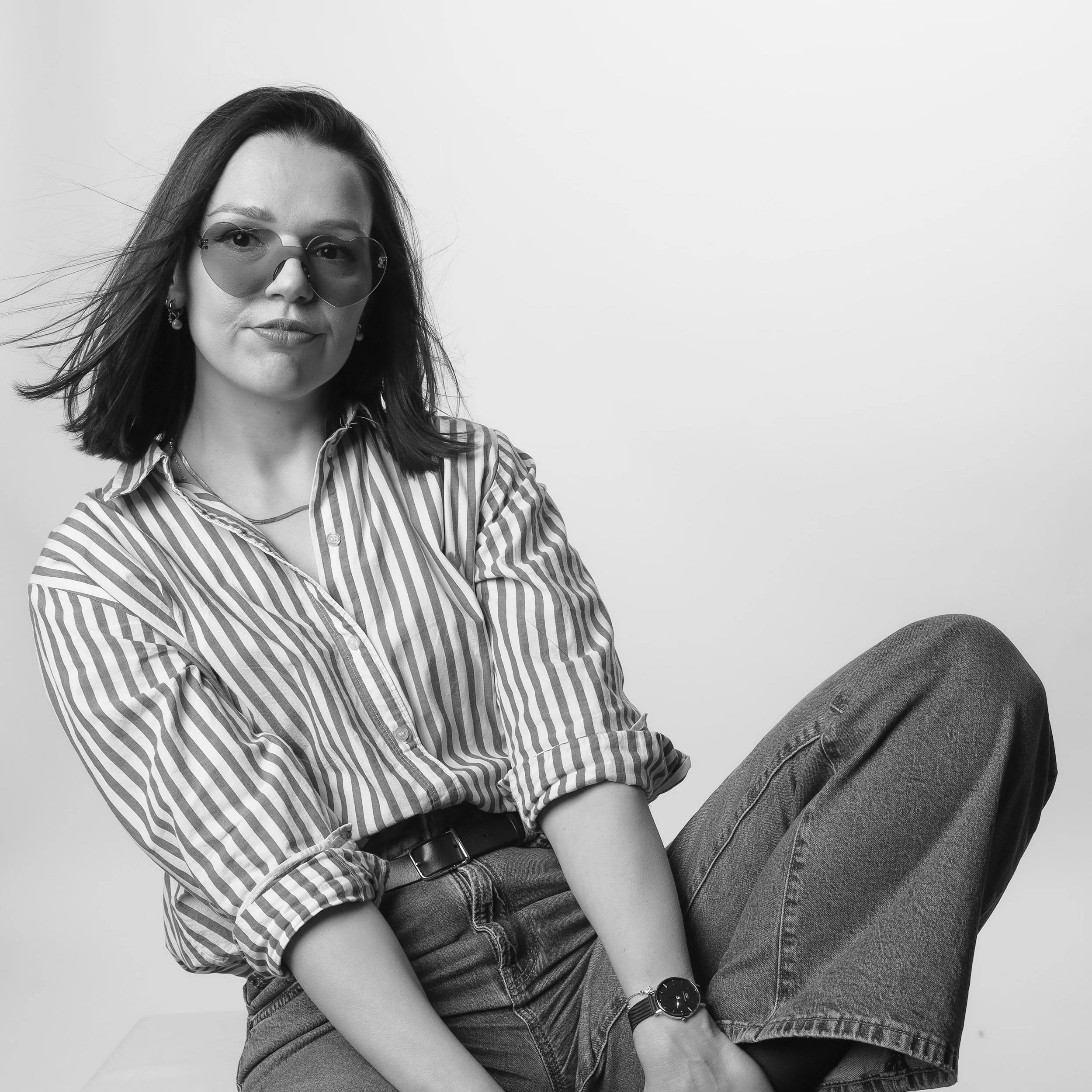 A woman with shoulder-length hair wearing sunglasses, a striped shirt, jeans, and a watch, sitting with one leg crossed over the other against a plain background.